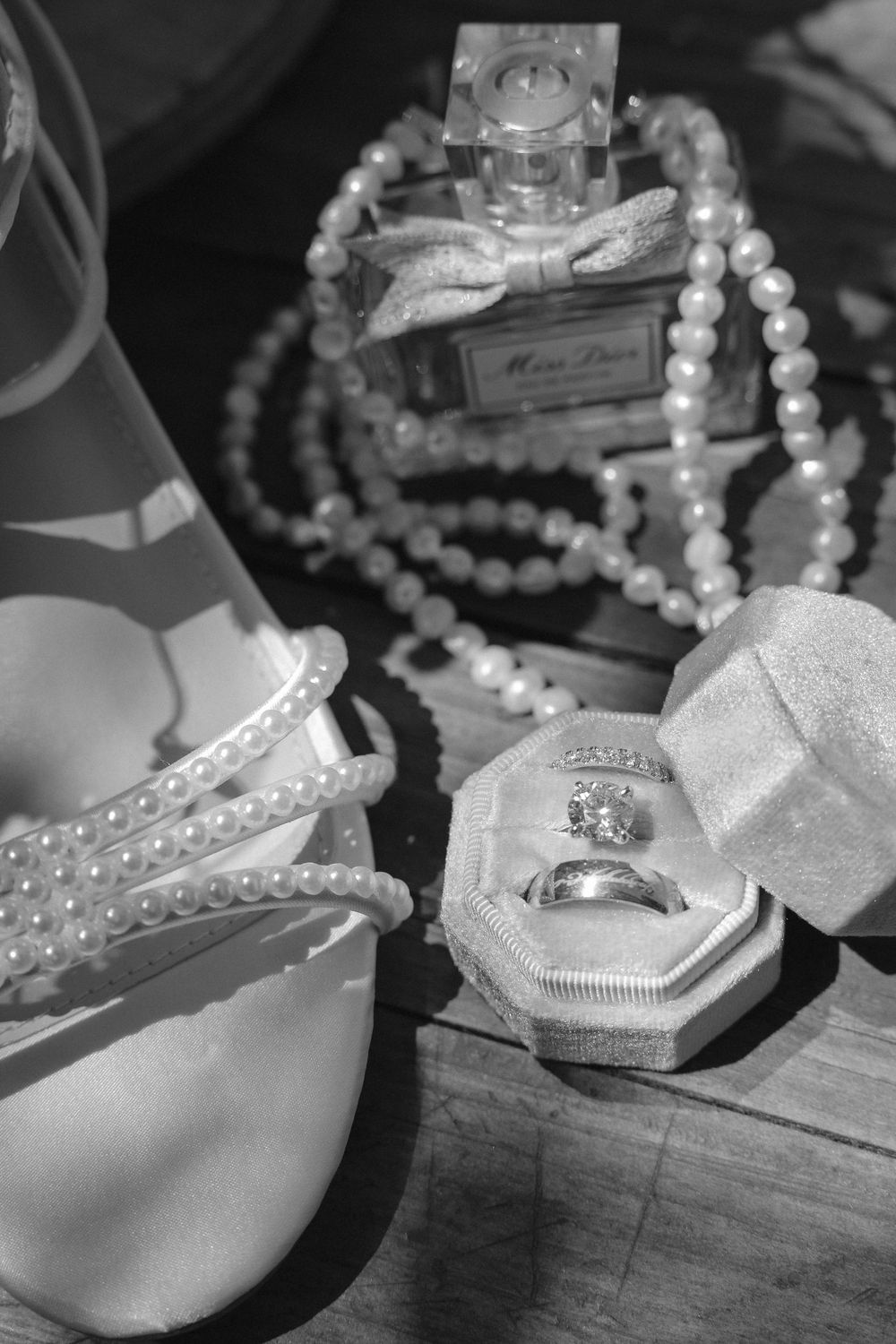 Black and white wedding details with pearl-strapped heels, a Miss Dior perfume bottle, a pearl necklace, and rings in a velvet ring box.