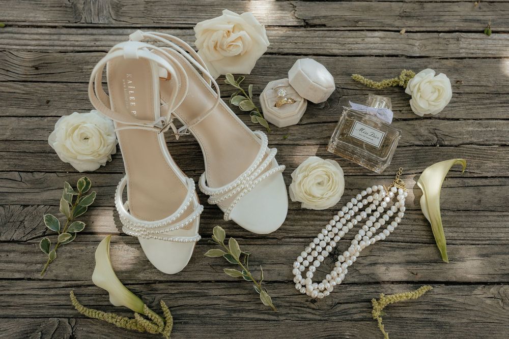 White strappy bridal sandals with pearl details, a pearl necklace, perfume bottle and cream roses arranged on rustic wooden planks.