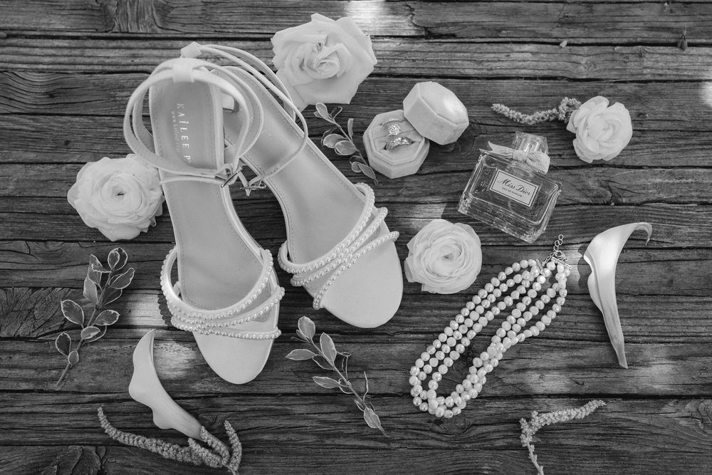 Black and white flat lay of pearl-embellished bridal sandals with roses, a pearl necklace, perfume bottle and compact on rustic wood.