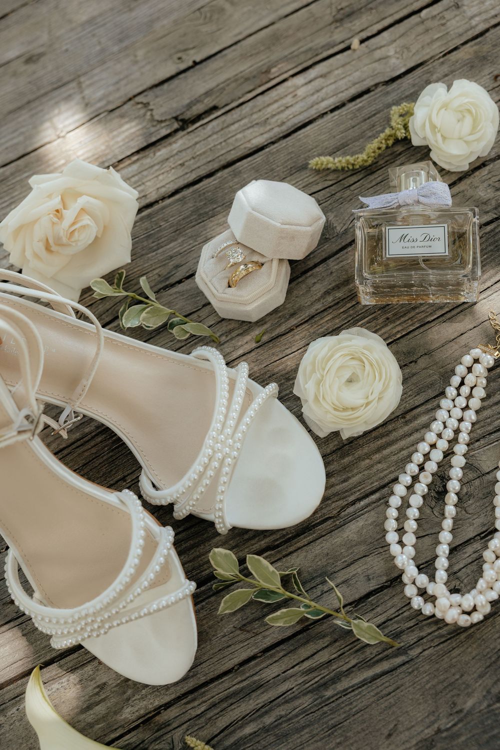 Wedding flat lay with pearl-strap ivory heeled sandals, cream roses, a ring box with a ring, Miss Dior perfume and a pearl necklace on wood.