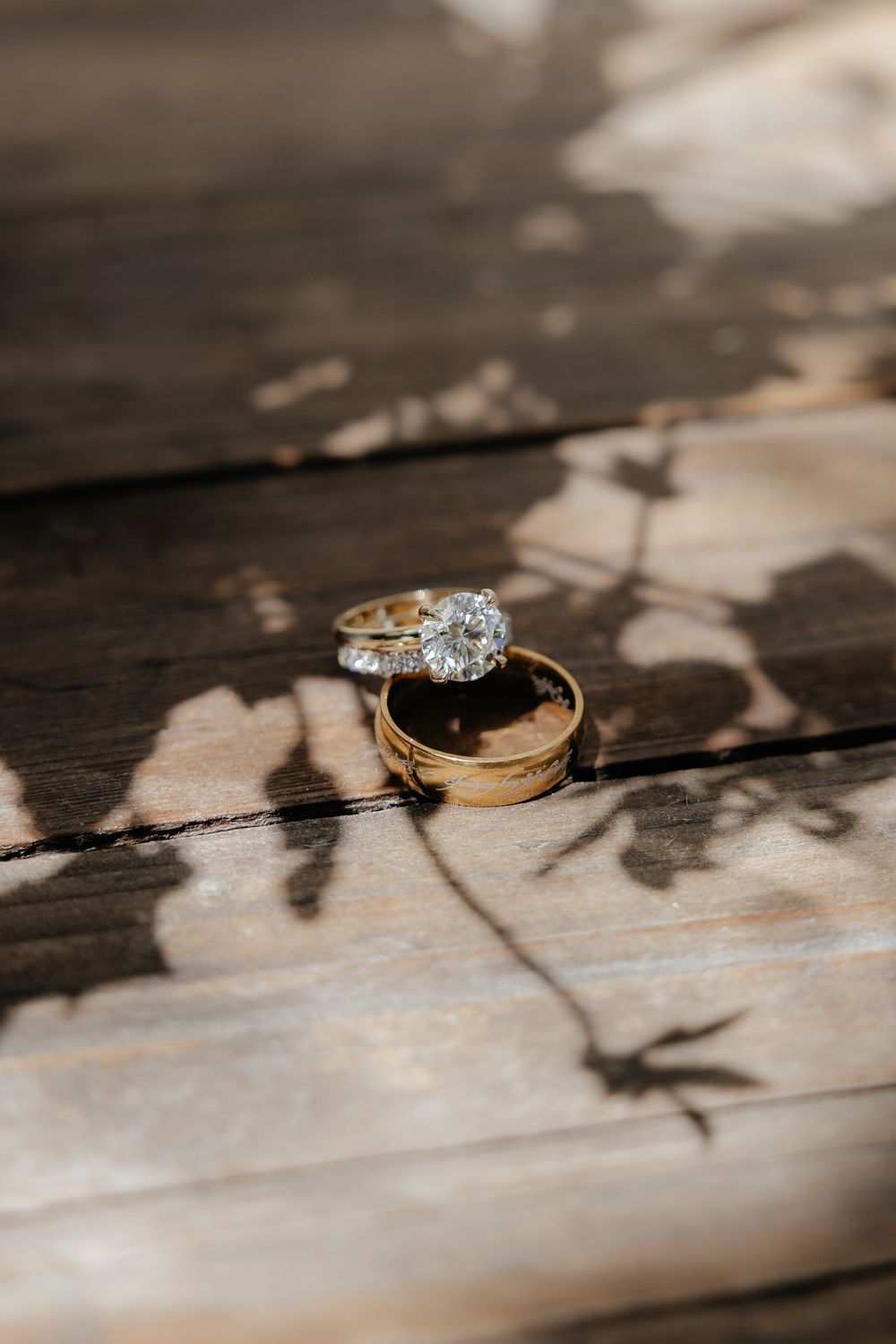 Diamond engagement ring and gold wedding band resting on weathered wooden planks with dappled leaf shadows.