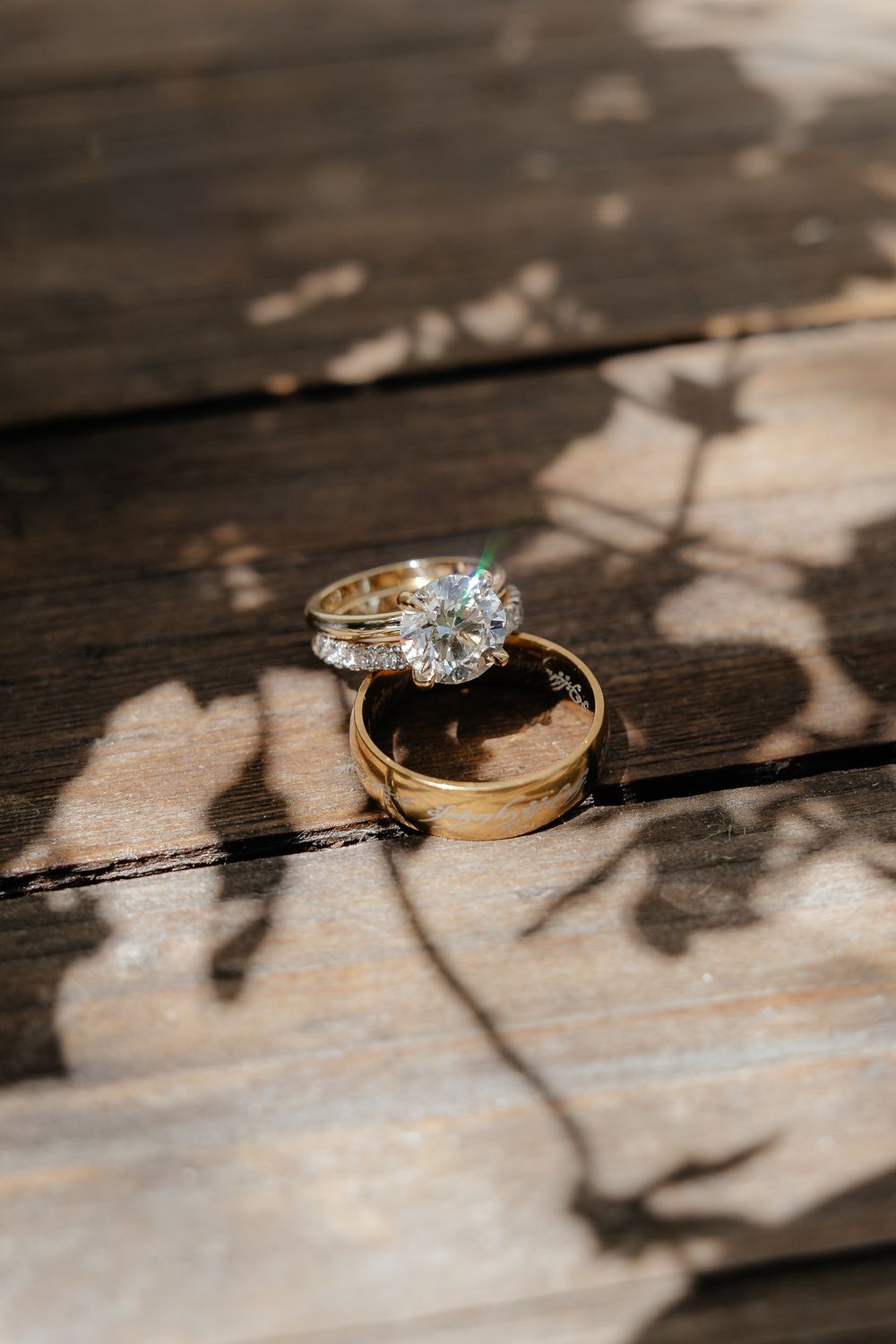 Diamond engagement ring and gold wedding band resting on a rustic wooden surface with dappled leaf shadows.