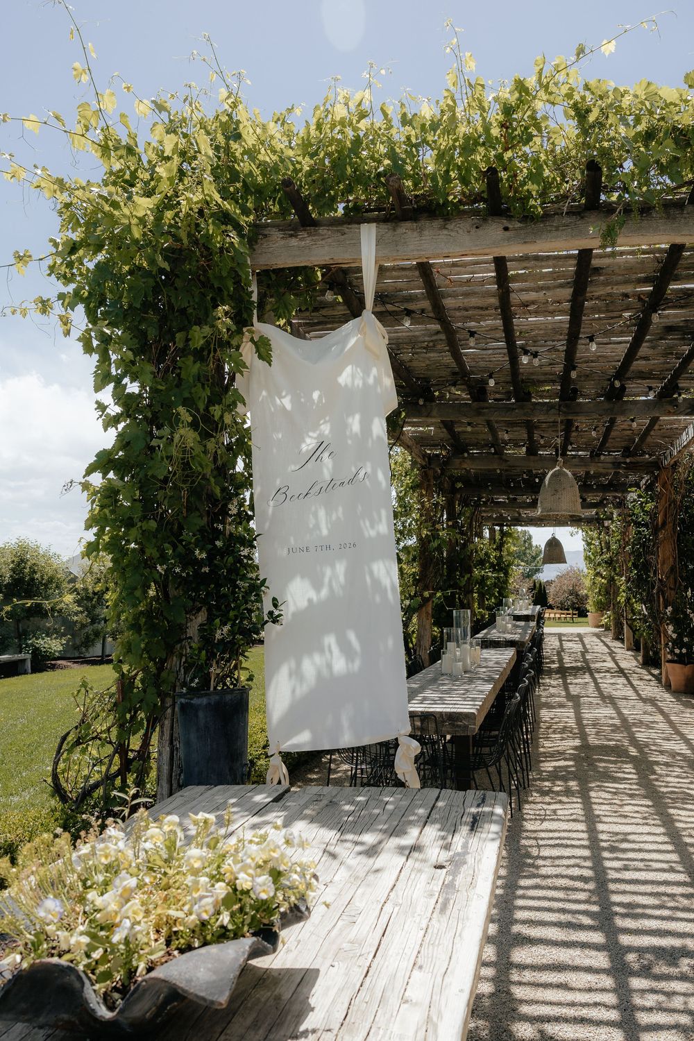Rustic wooden pergola draped in green vines over a long banquet table, with a white welcome banner hanging in a sunny garden venue.