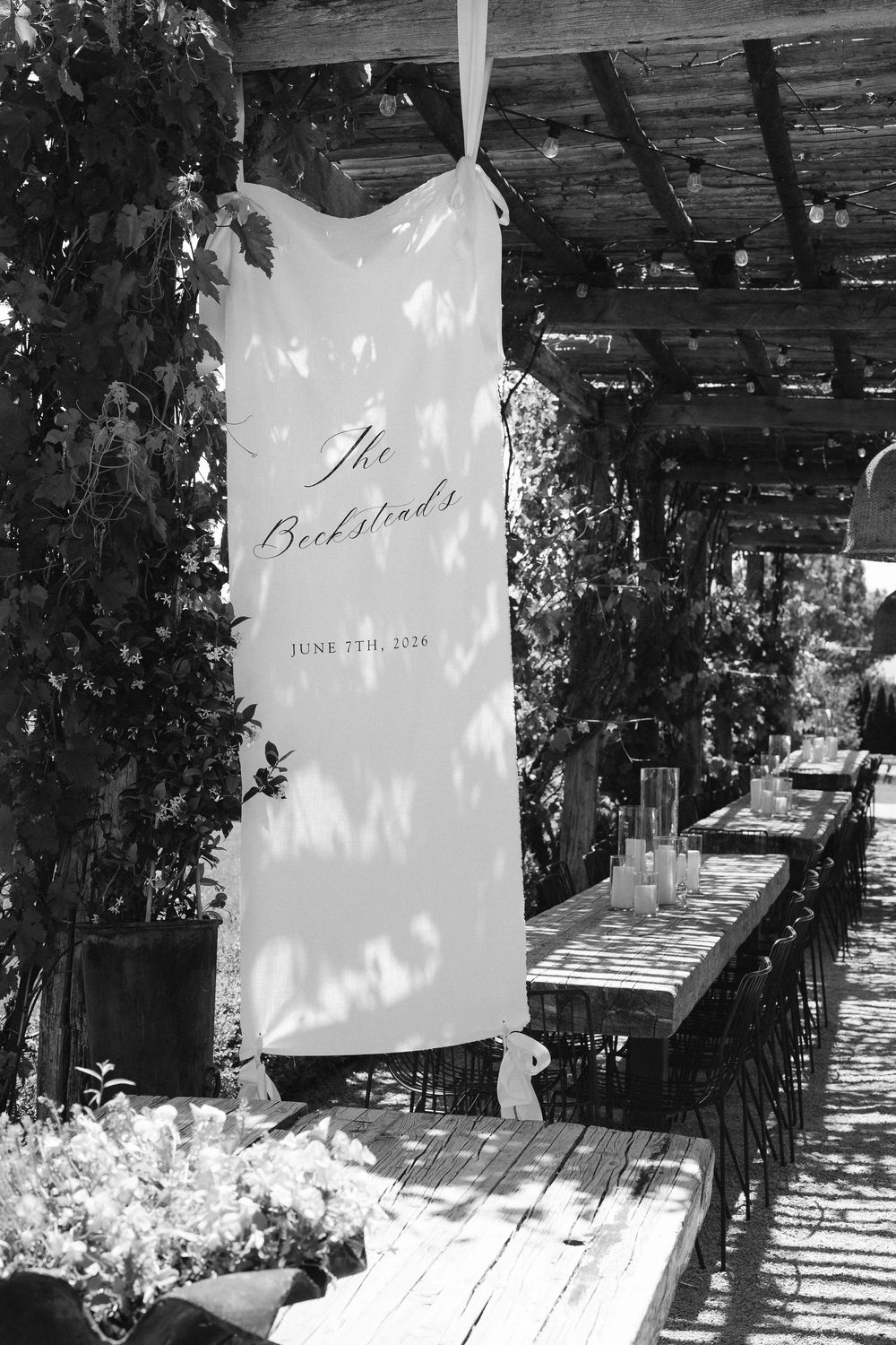 Black and white outdoor wedding reception with long wooden tables under a pergola and a hanging welcome banner.