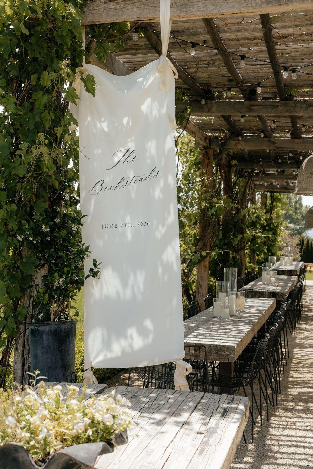 Rustic wooden tables and black chairs under a vine-covered pergola, with a white welcome banner reading The Beckstead's dated June 2026.