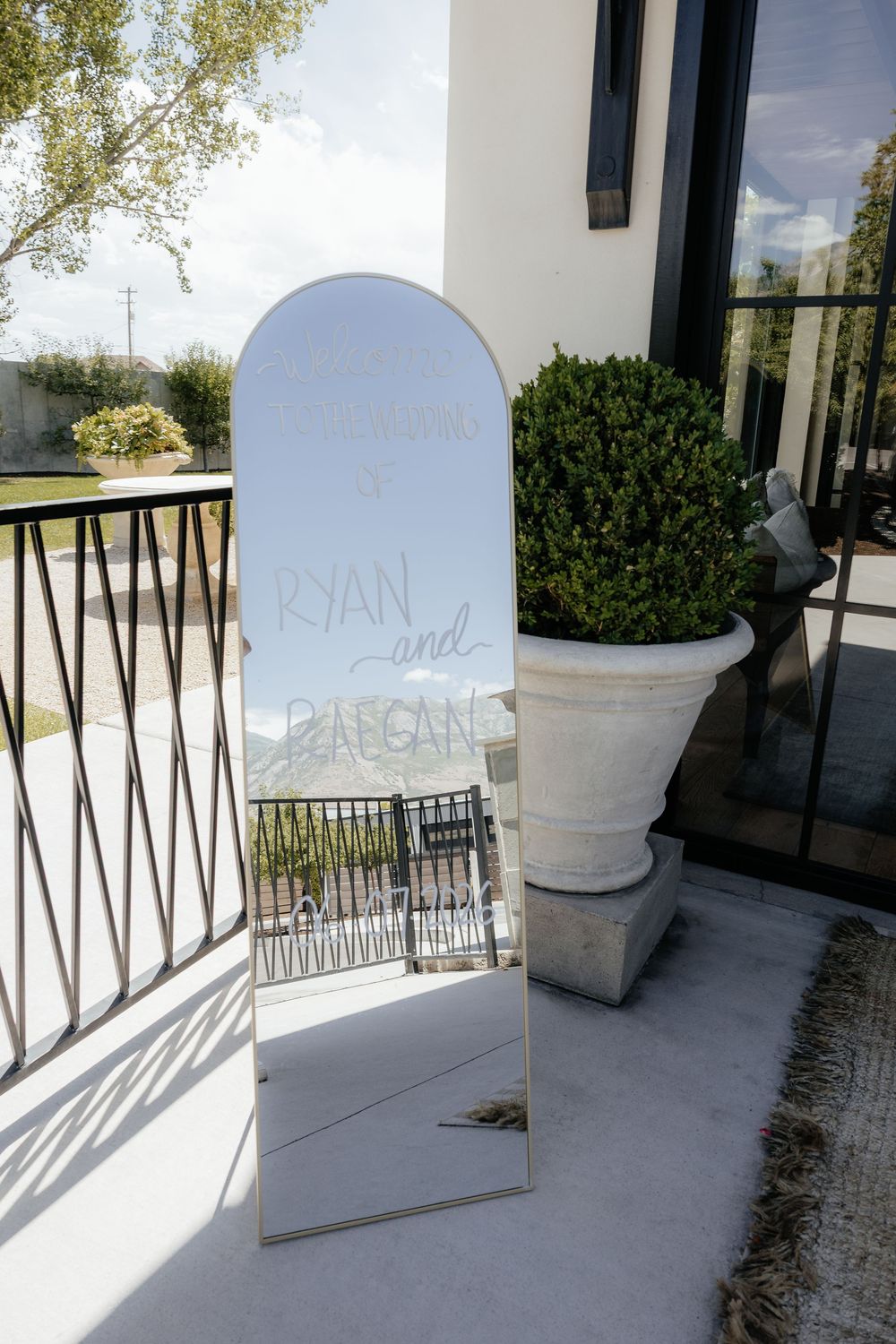 Arched standing mirror with handwritten Ryan and welcome lettering beside a potted boxwood shrub at a sunny wedding venue entrance.
