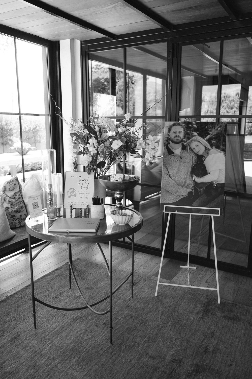 Black and white wedding welcome table with floral arrangement, guest book and framed couple portrait on an easel in a sunlit glass room.