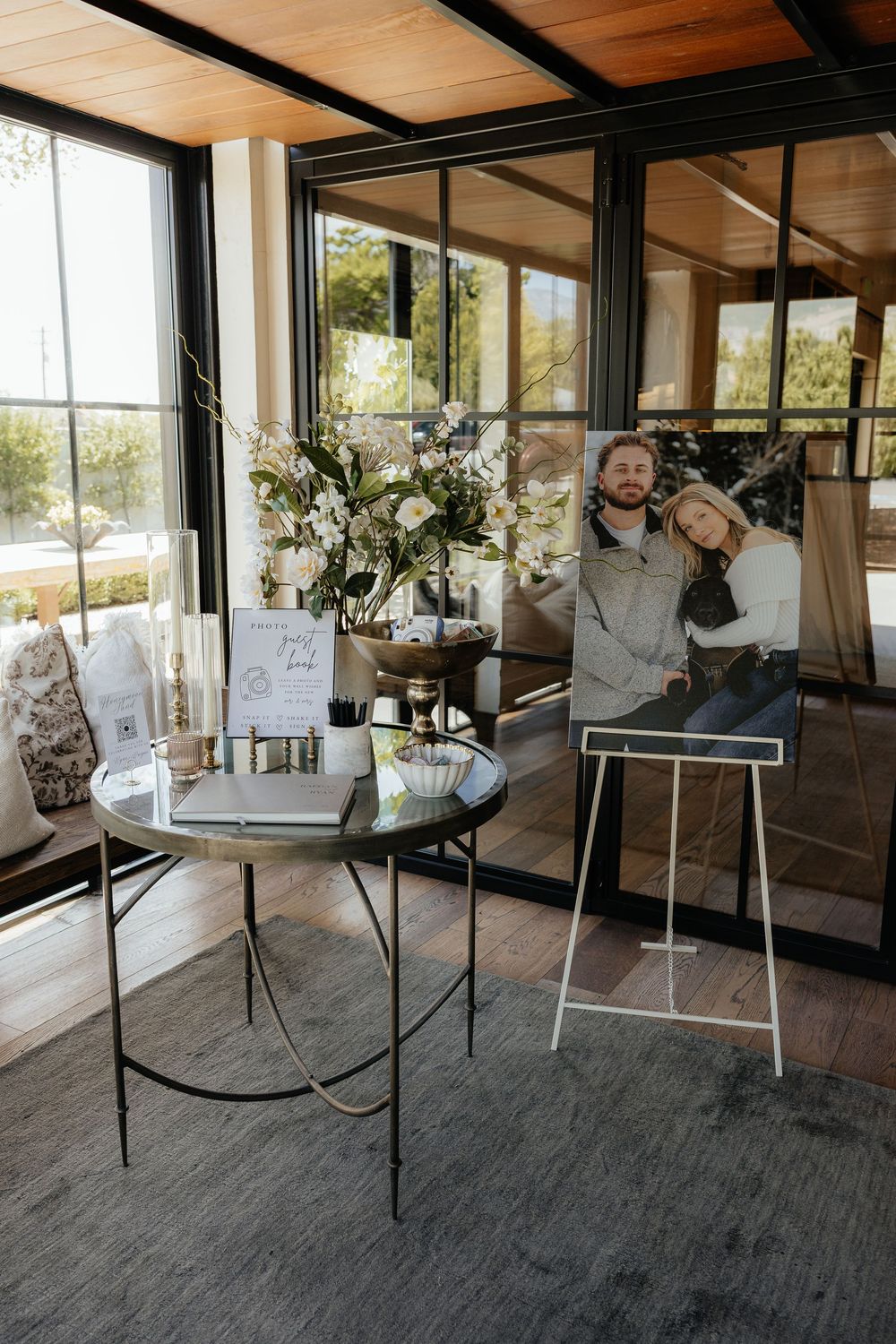 Wedding welcome table with floral arrangement, guest book and framed couple portrait on an easel in a bright glass-walled venue.