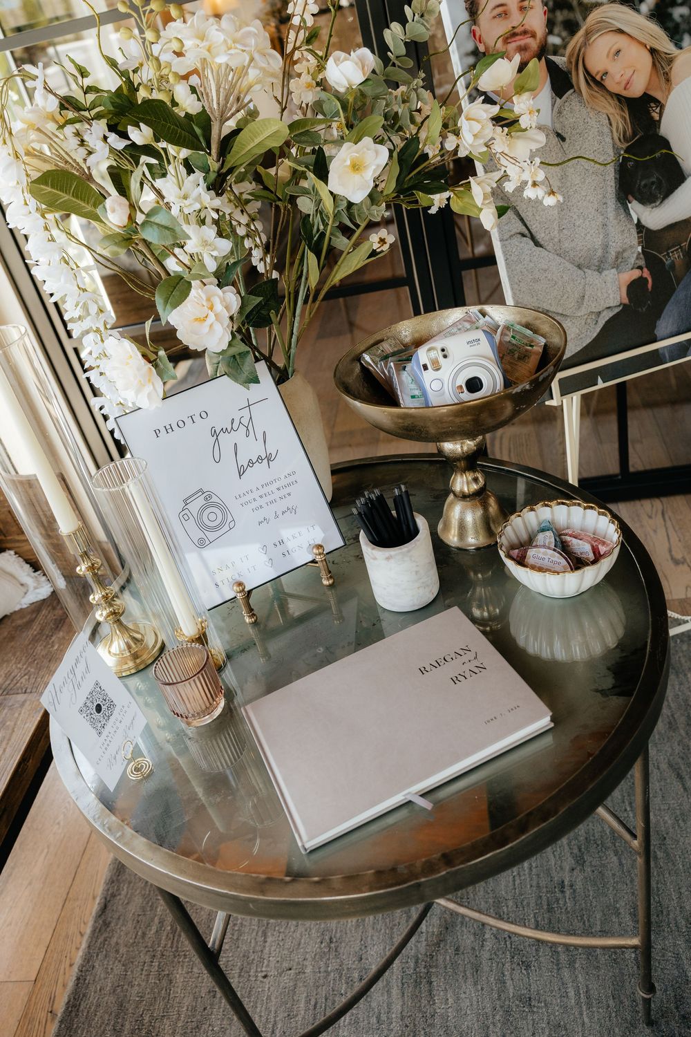 Wedding photo guest book table with an instant camera, white floral arrangement, pens, candles and a sign on a round glass-top table.