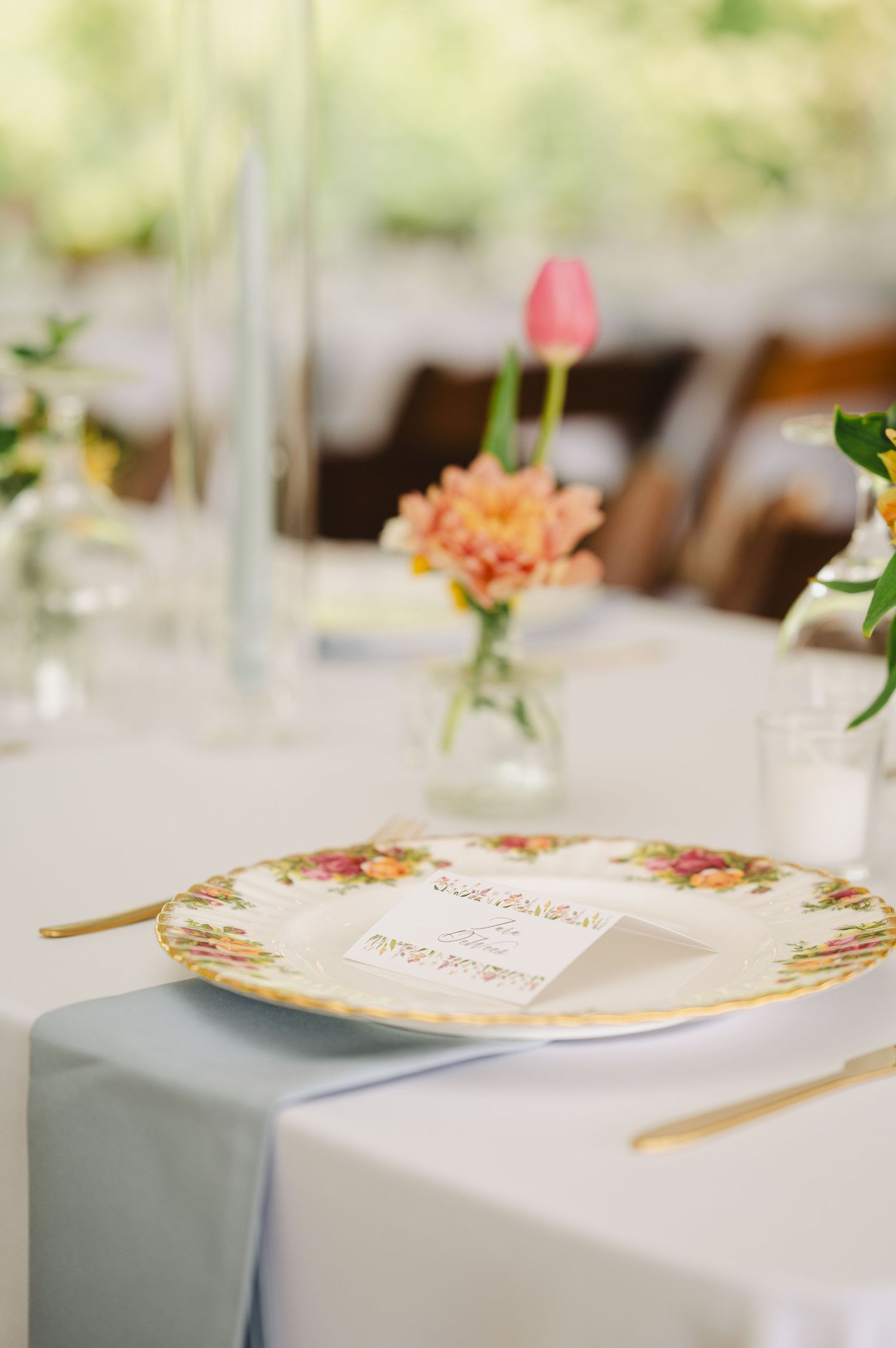 Elegant wedding table setting with floral china plate, gold cutlery, menu card, and pink flowers in bud vases.