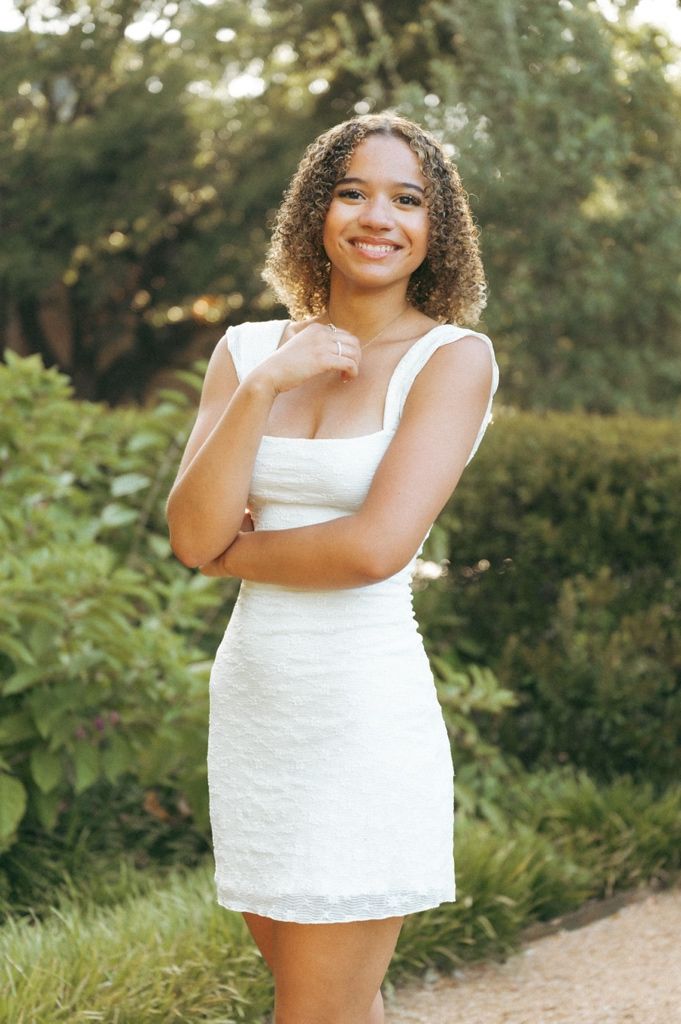Young woman in white mini dress smiling outdoors, posing gracefully among lush green garden hedges.