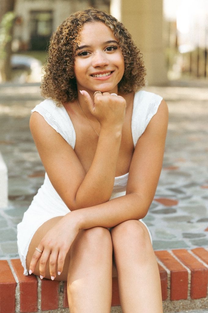 A smiling young woman with curly hair sits on brick steps wearing a white dress in an outdoor setting.
