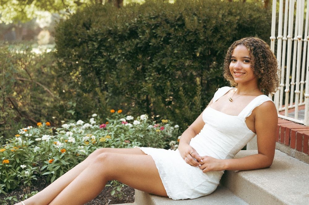 A smiling young woman in a white dress sits on stone steps surrounded by lush greenery and colorful flowers outdoors.