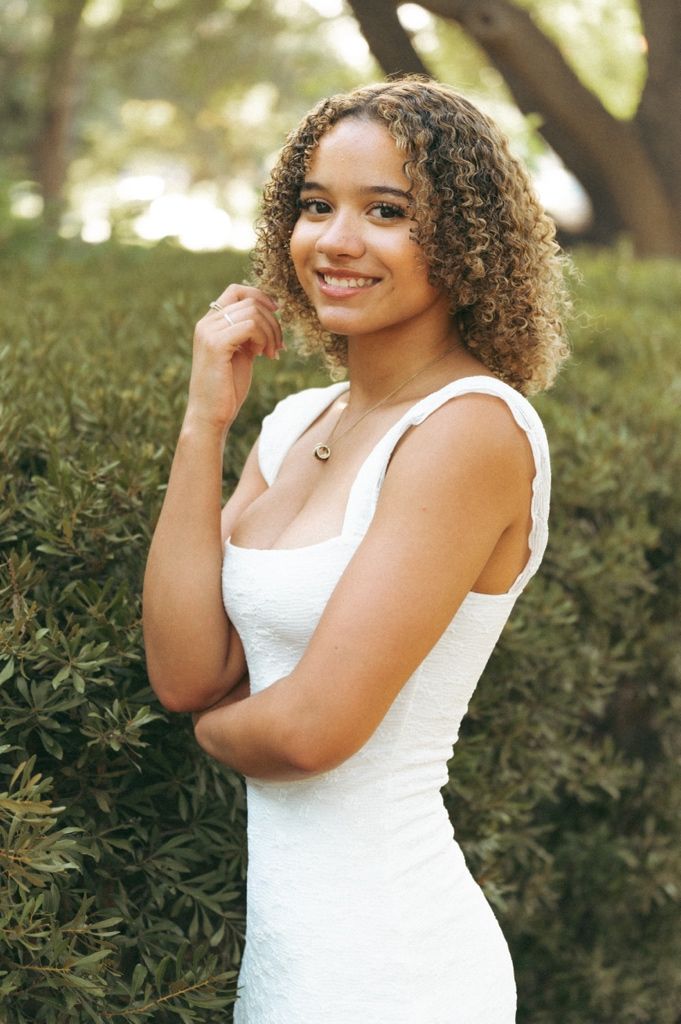 Young woman with curly hair smiling in a white dress, posing outdoors near green hedges in natural light.