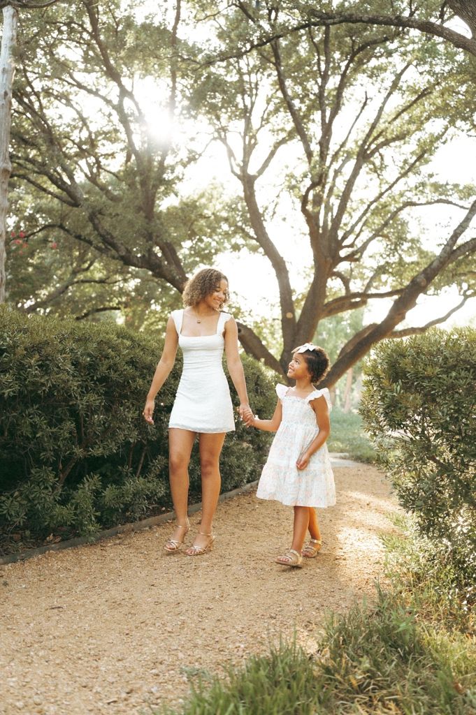 Woman and young girl in white dresses holding hands walking on a garden path surrounded by lush greenery.