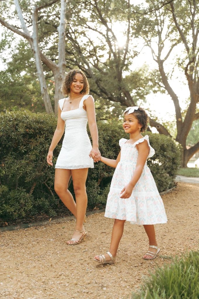 A woman and young girl in matching white dresses hold hands while walking along a tree-lined path outdoors.
