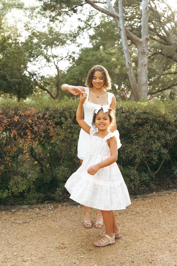 A woman and young girl in matching white dresses pose together on a garden path surrounded by lush green hedges.