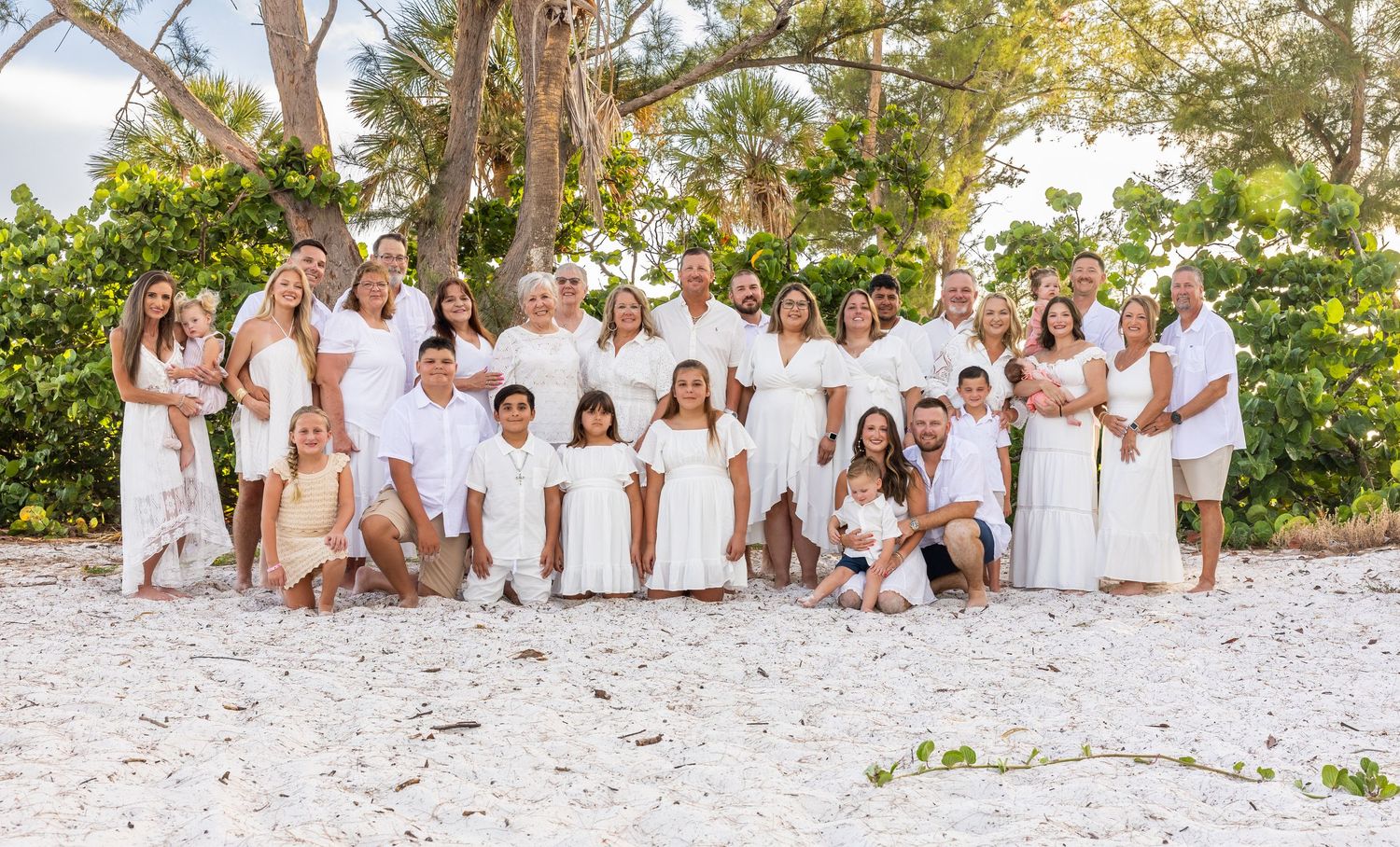 Large family group photo on sandy beach, all wearing white outfits, posing under tropical trees at sunset.