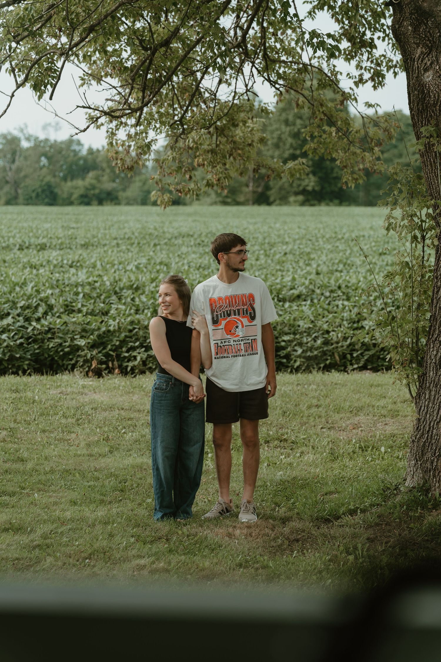 A couple holds hands in a lush green field, standing under a tree on a warm summer day.