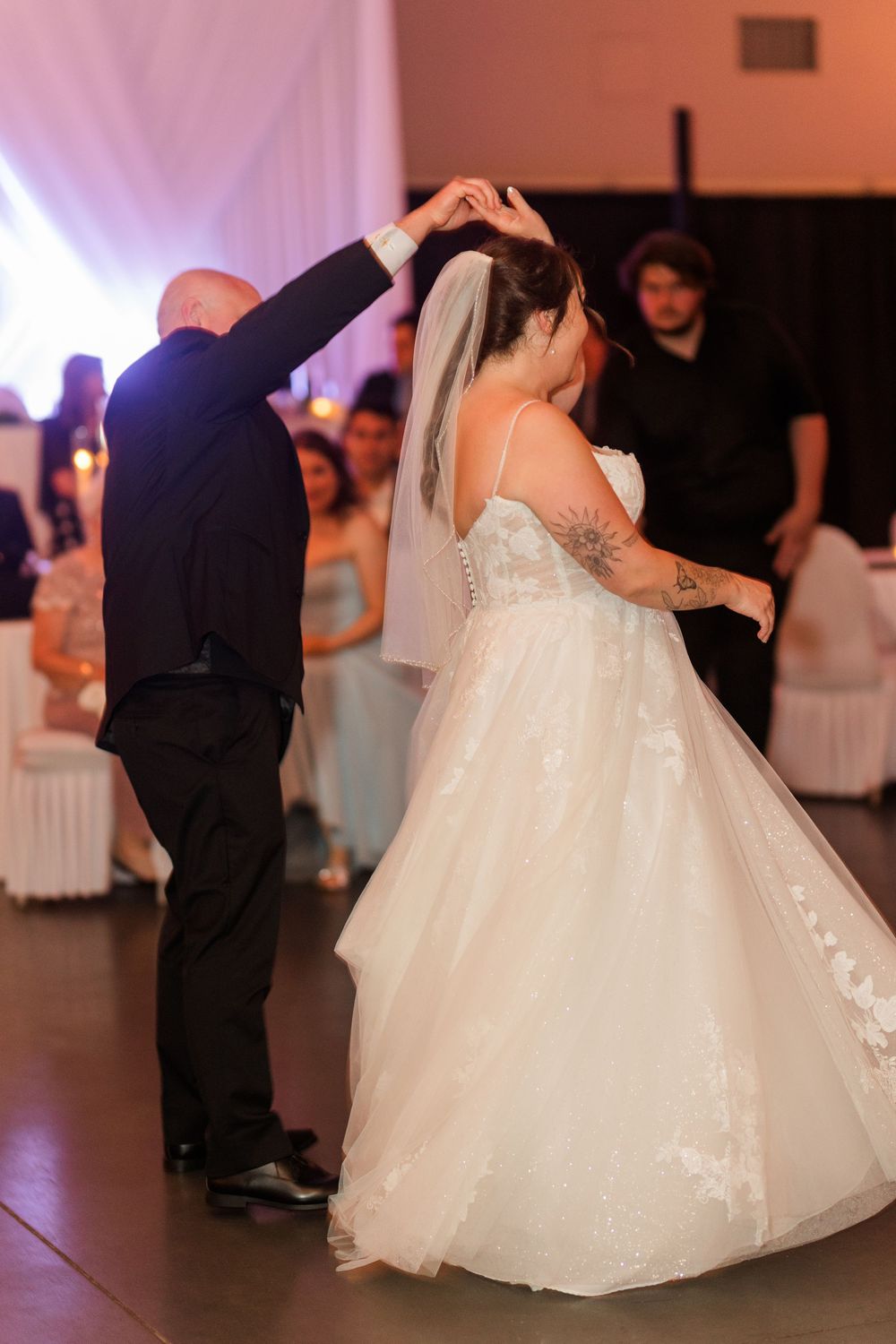 Bride in white ballgown spins during first dance with bald groom in black suit at wedding reception.