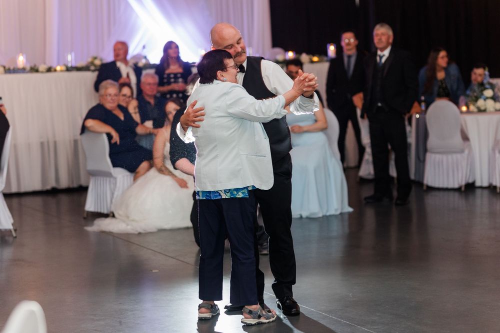 Groom shares a tender first dance with his mother at a wedding reception, guests watching from tables in the background.
