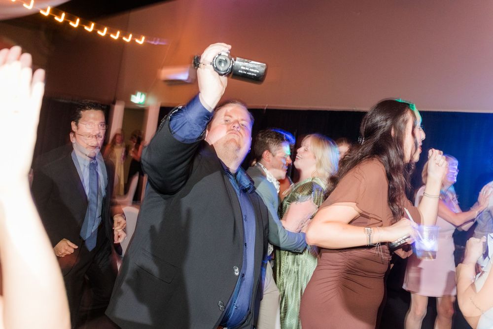 Man holding camcorder overhead while dancing at a lively indoor party with guests celebrating in background.