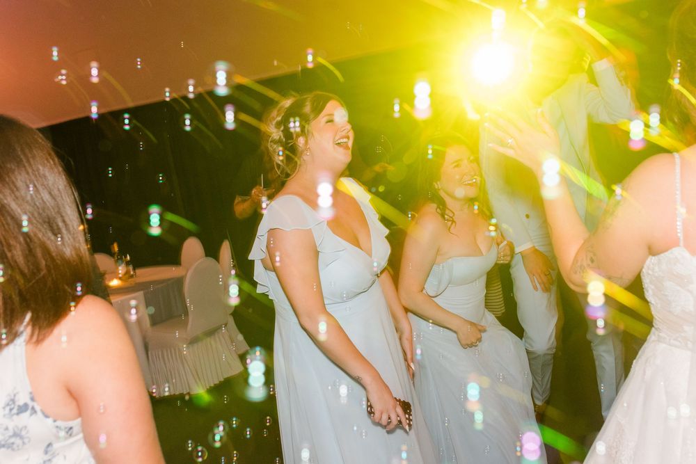 Bridesmaids in light blue dresses dancing joyfully on a wedding reception dance floor with bubbles and colorful lights.
