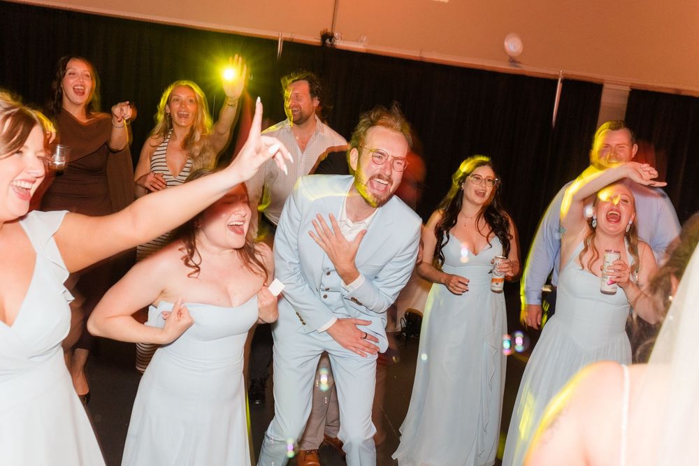 Wedding guests in light blue bridesmaid dresses dance and celebrate joyfully at a lively indoor wedding reception.