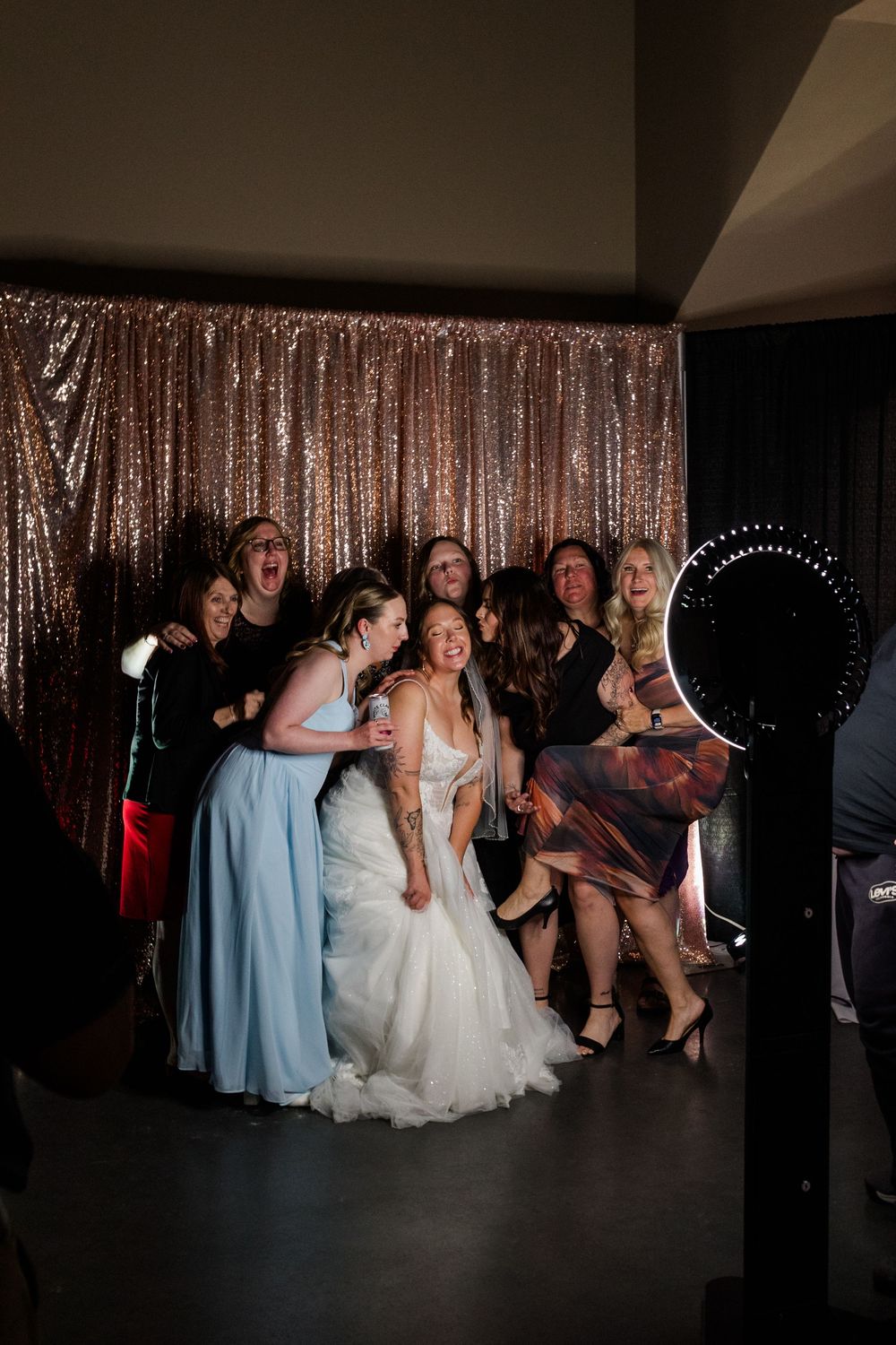 Bride in white gown laughs with bridesmaids and guests in front of gold sequin backdrop at wedding photo booth.