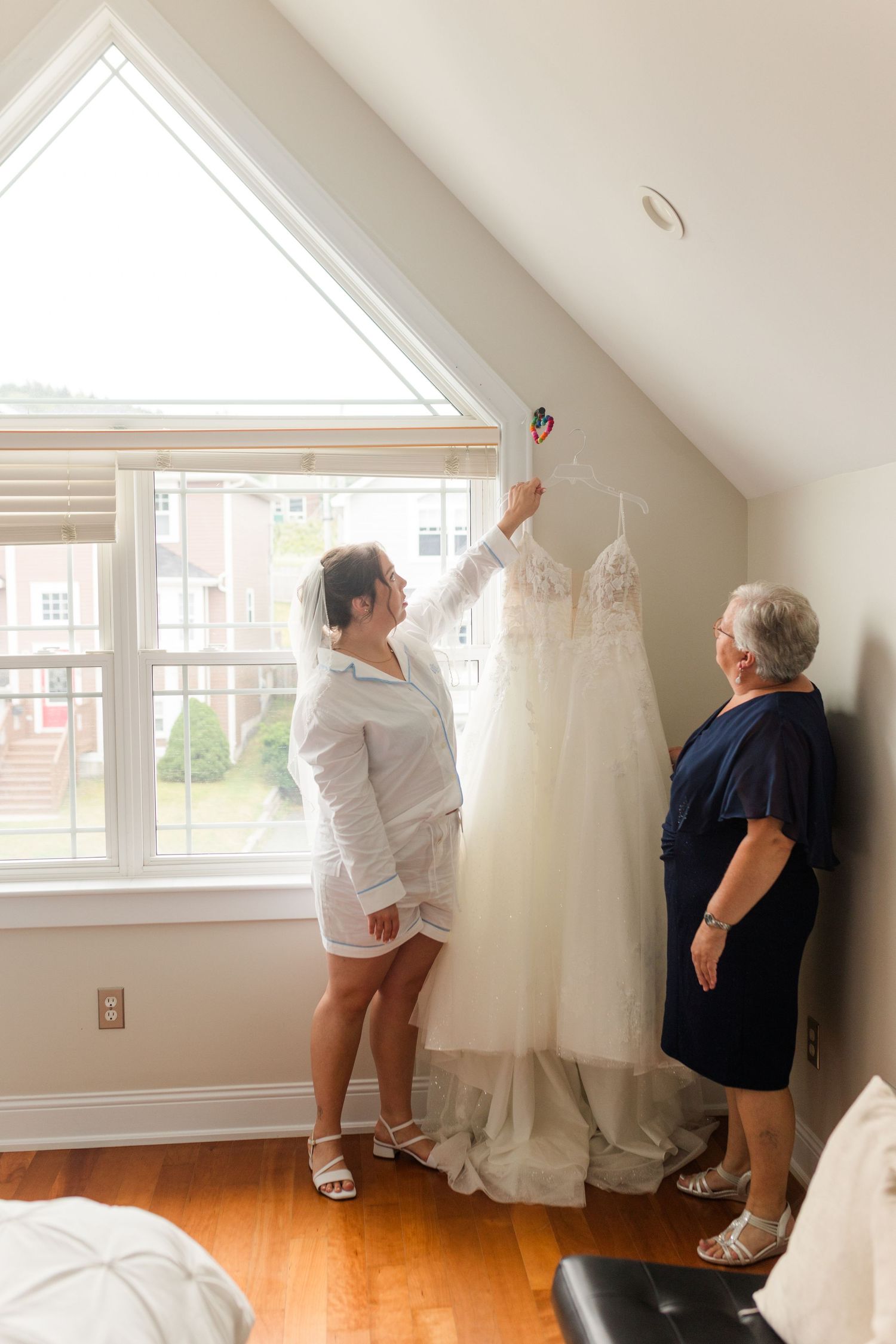 Bride and older woman hanging a white wedding gown near a large triangular window in a bright room.
