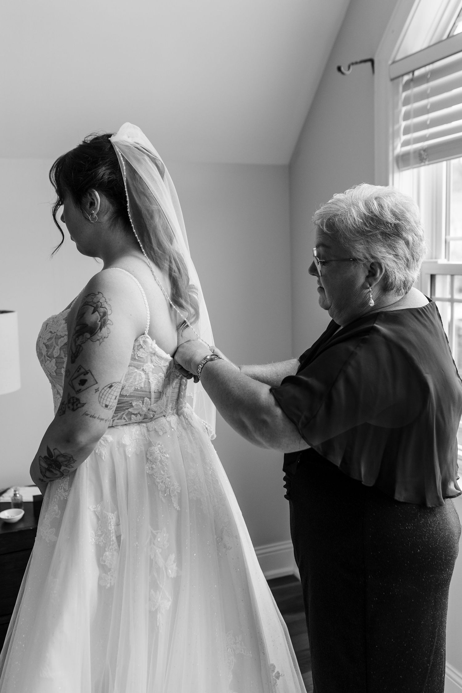 Black and white photo of elderly woman helping bride with wedding dress and veil before ceremony.
