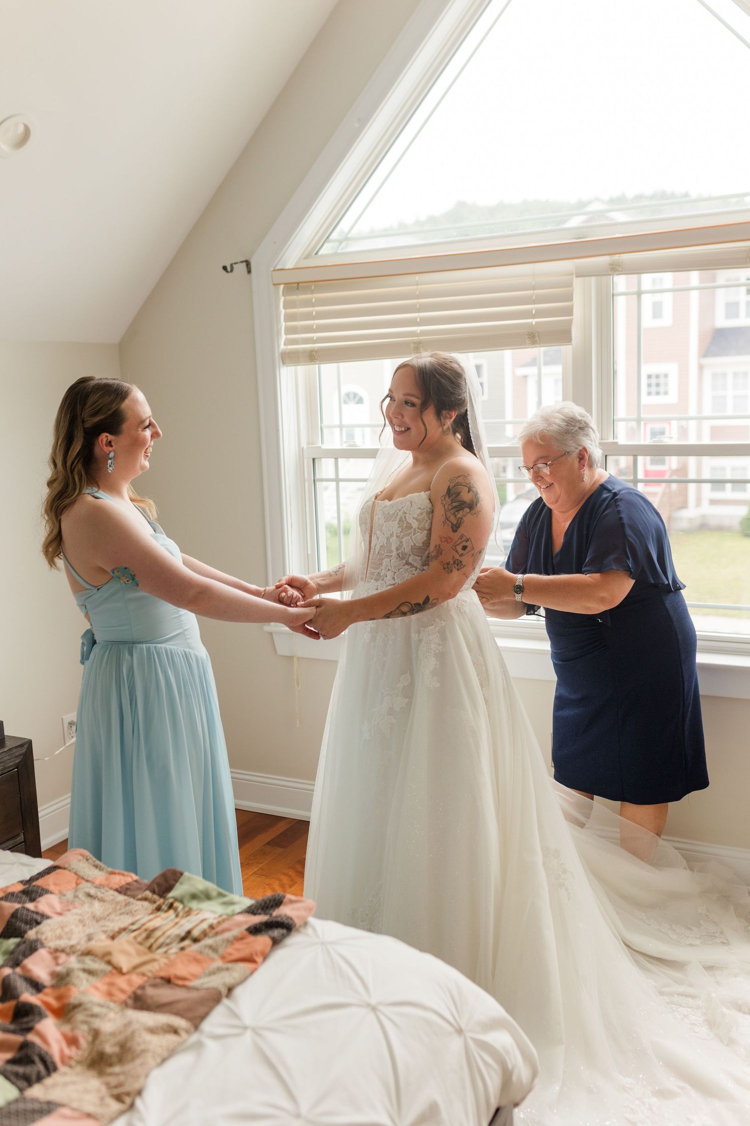 Bride in white gown holds hands with bridesmaid in blue dress while older woman adjusts the wedding dress train indoors.