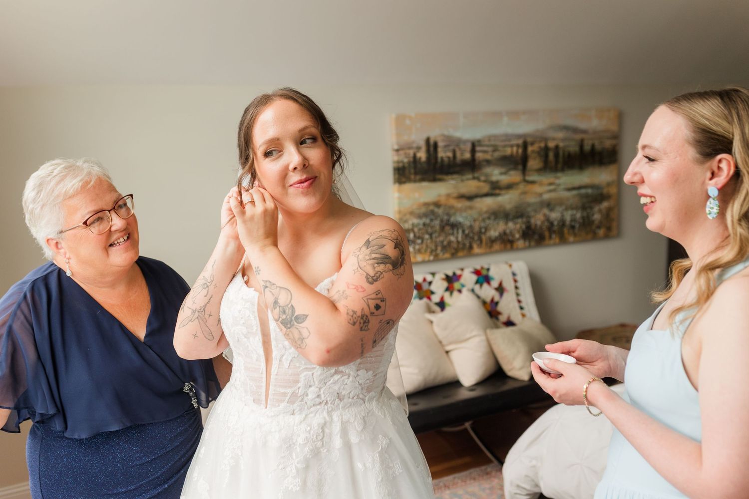 Bride in white lace gown putting on earrings assisted by older woman and bridesmaid in light blue dress.