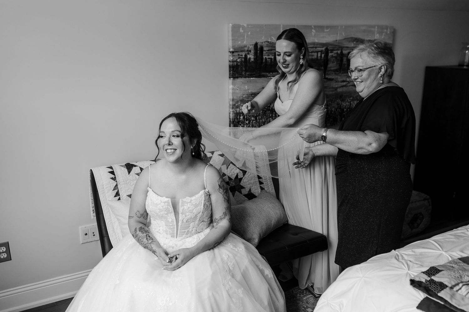 Black and white photo of a bride in a wedding gown being helped with her veil by two women in a hotel room.