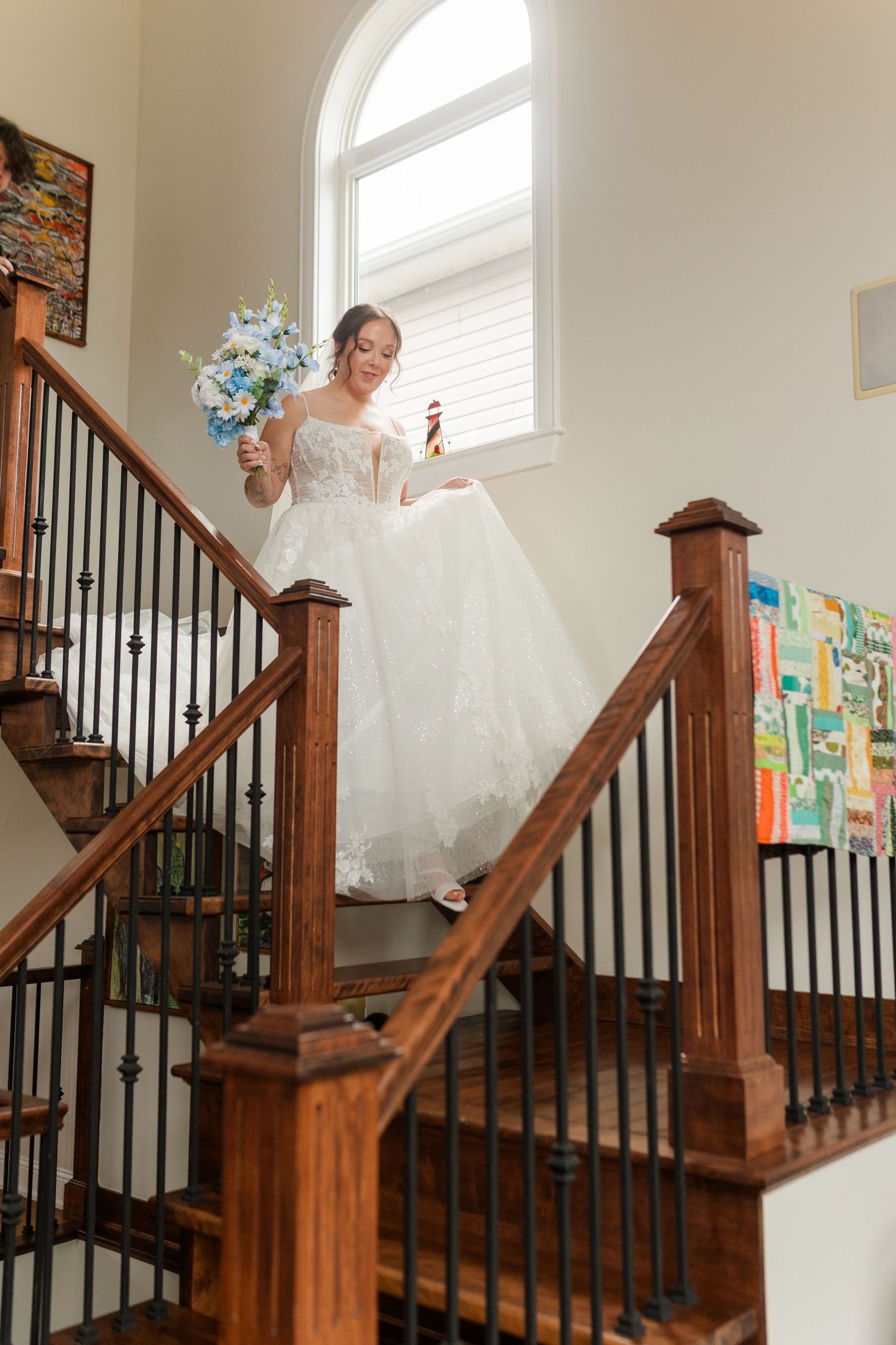 Bride in white lace gown descending wooden staircase holding blue and white bouquet, elegant indoor wedding portrait.