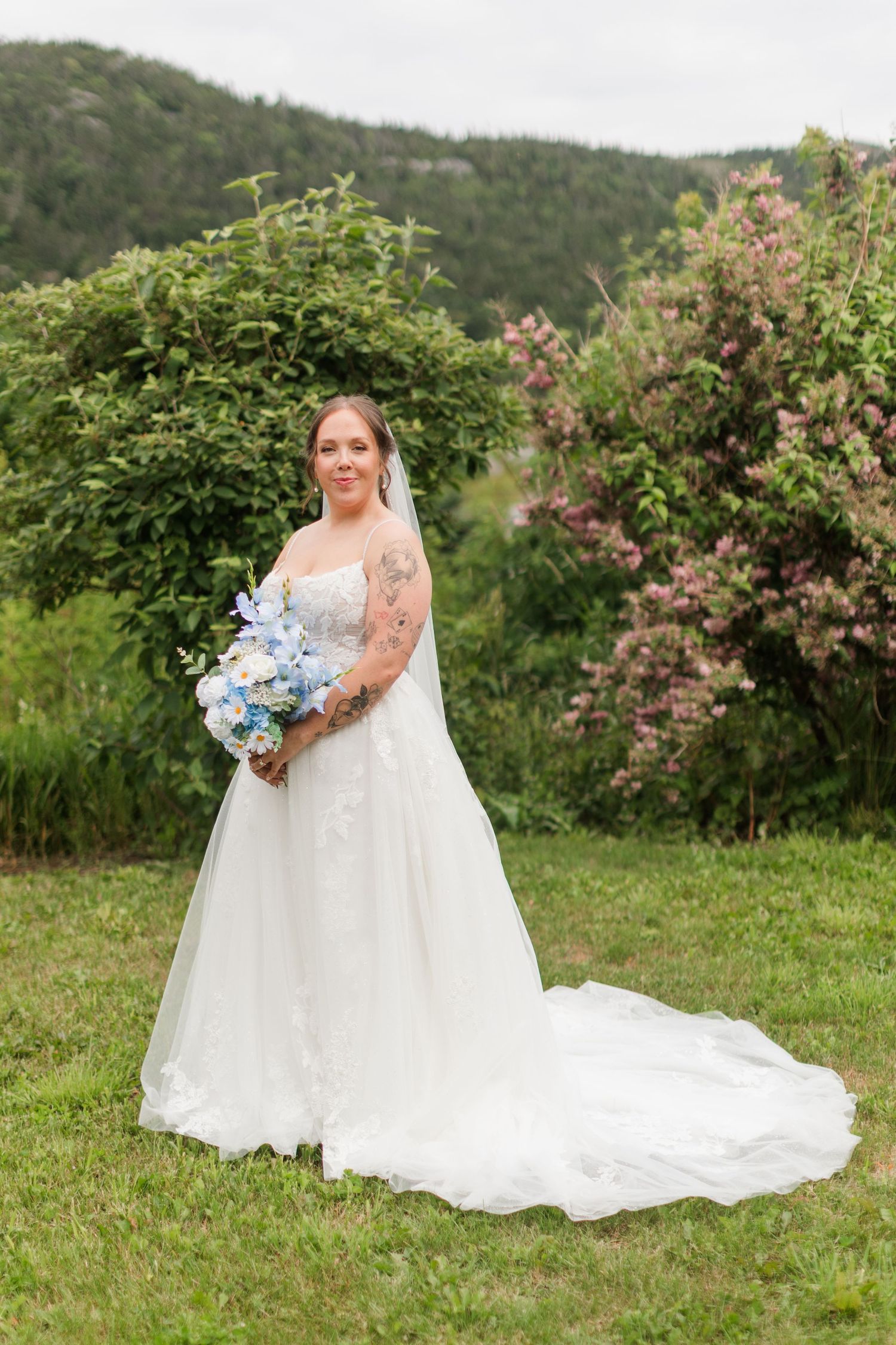 Bride in white lace ball gown holding blue floral bouquet, standing in lush garden with mountain backdrop.