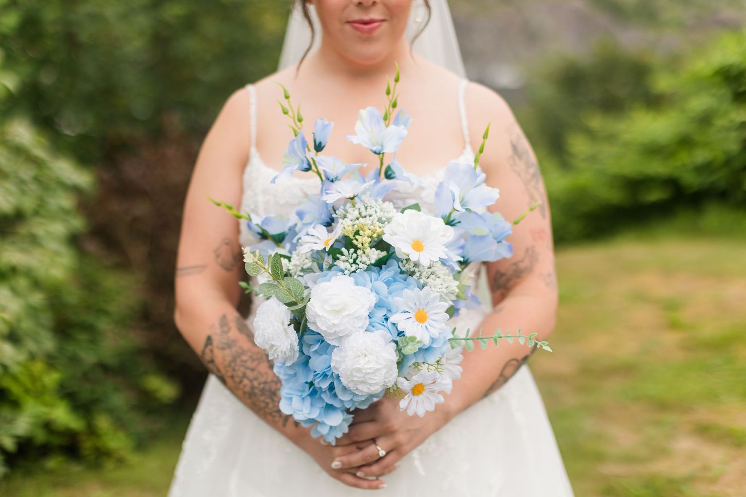 Bride in white gown holding a bouquet of blue hydrangeas and white flowers, with tattoos visible on her arms.