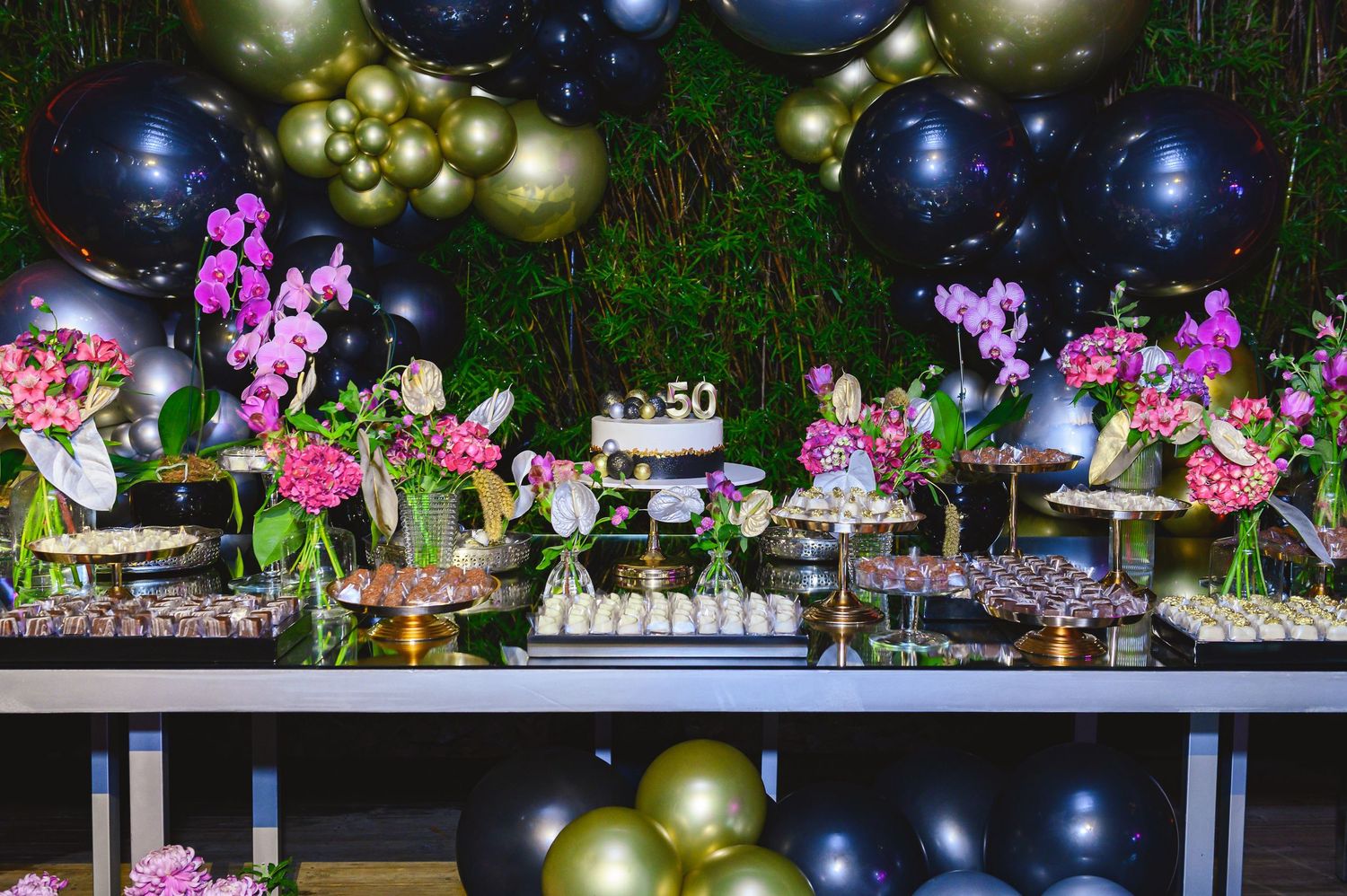 Elegant 50th birthday dessert table with black and gold balloons, pink orchids, and tiered cake stands.