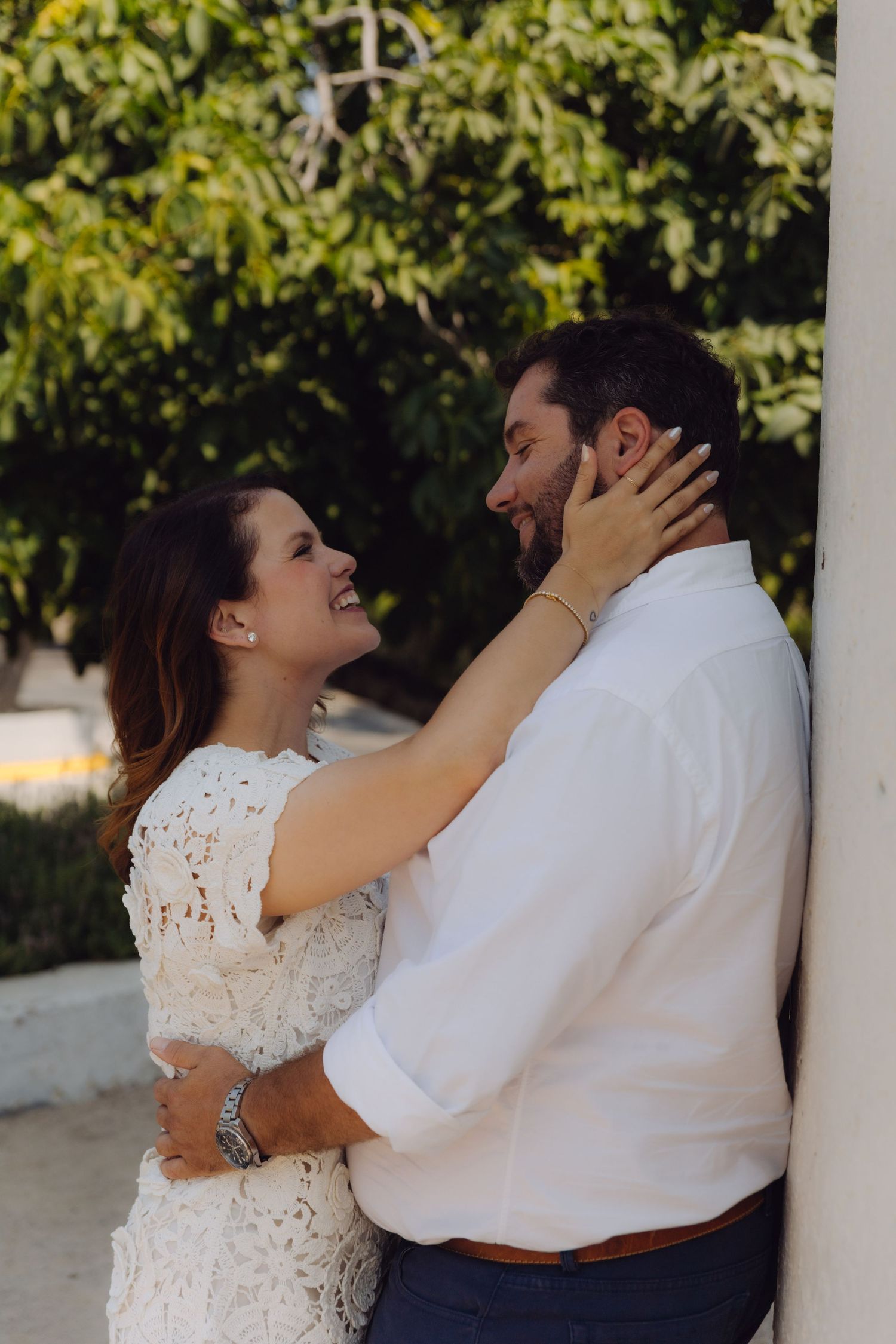 Laughing bride tenderly holding grooms face while embracing against a pillar outdoors.