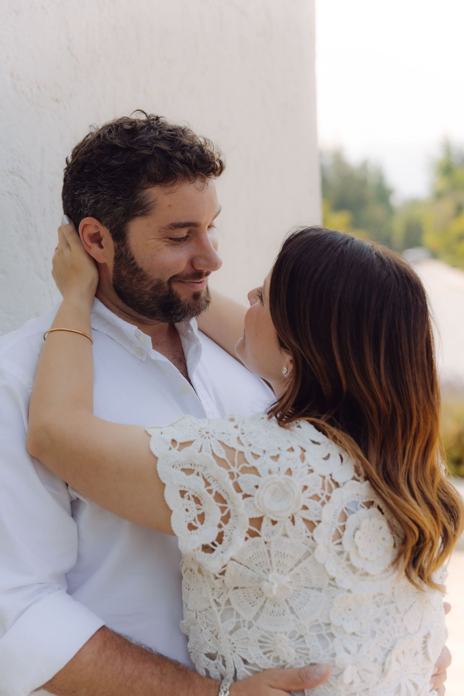 Romantic couple sharing an intimate moment, woman in white lace dress embracing bearded man in white shirt against a wall.