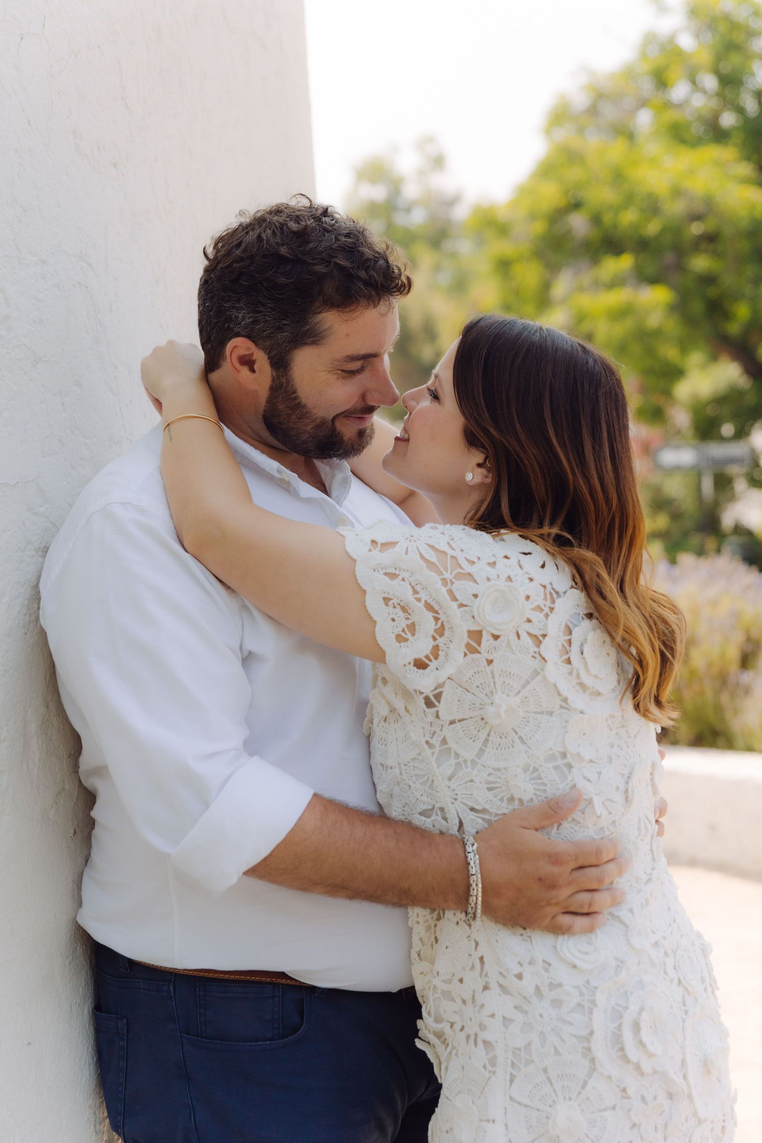 Couple almost kissing outdoors, woman in white lace dress and man in white shirt embracing against a light wall.