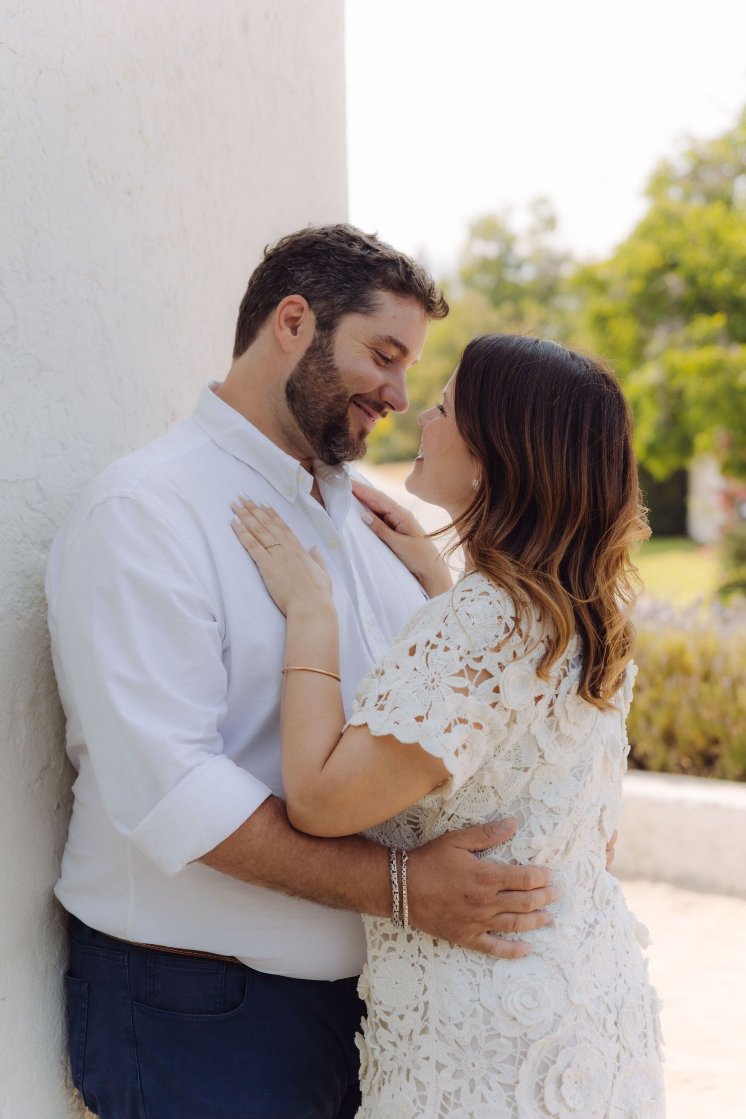 Loving couple gazing at each other, woman in white lace dress and bearded man in white shirt embracing outdoors.