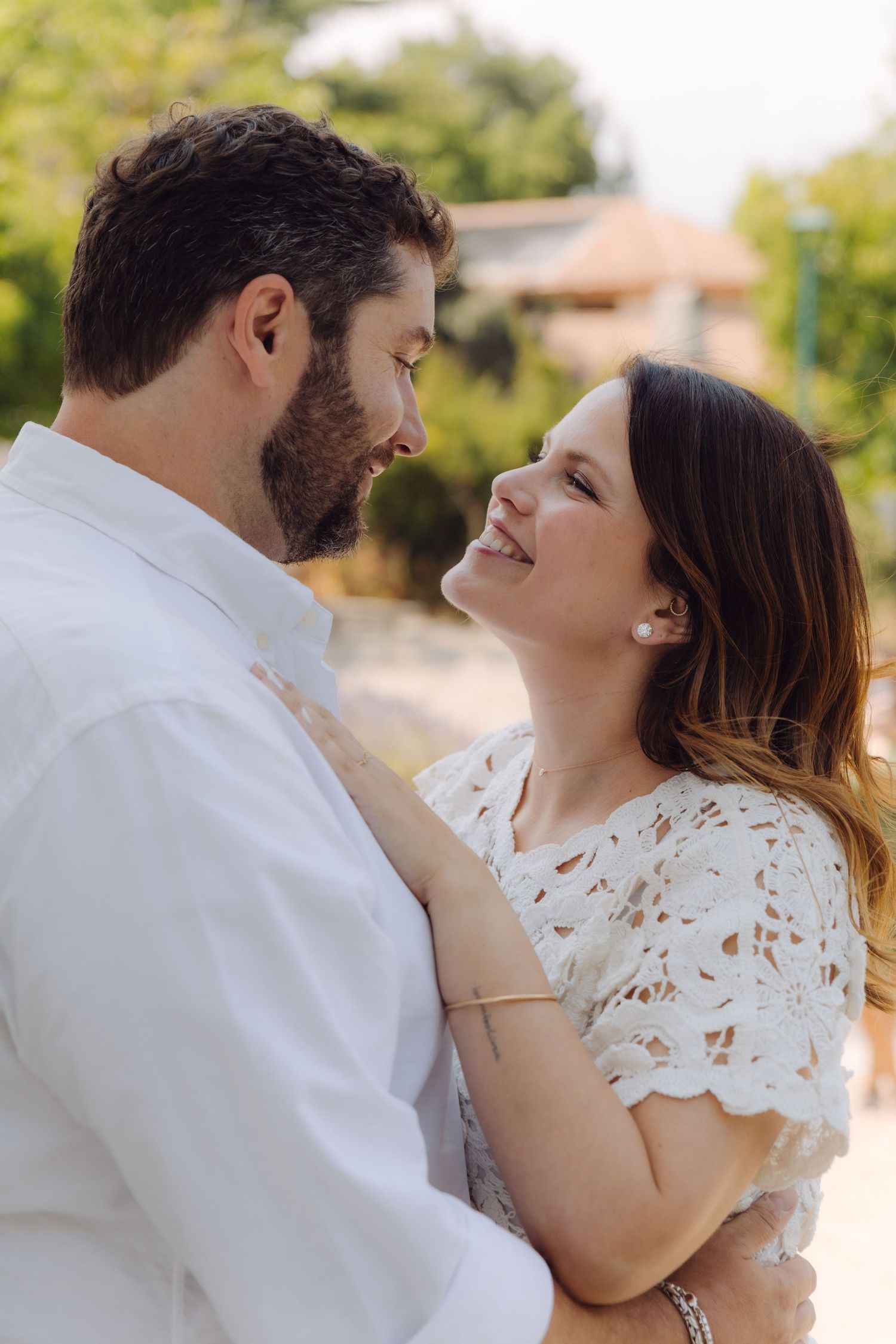 Happy woman in white lace dress smiling up at bearded man in white shirt during outdoor couples photo session.