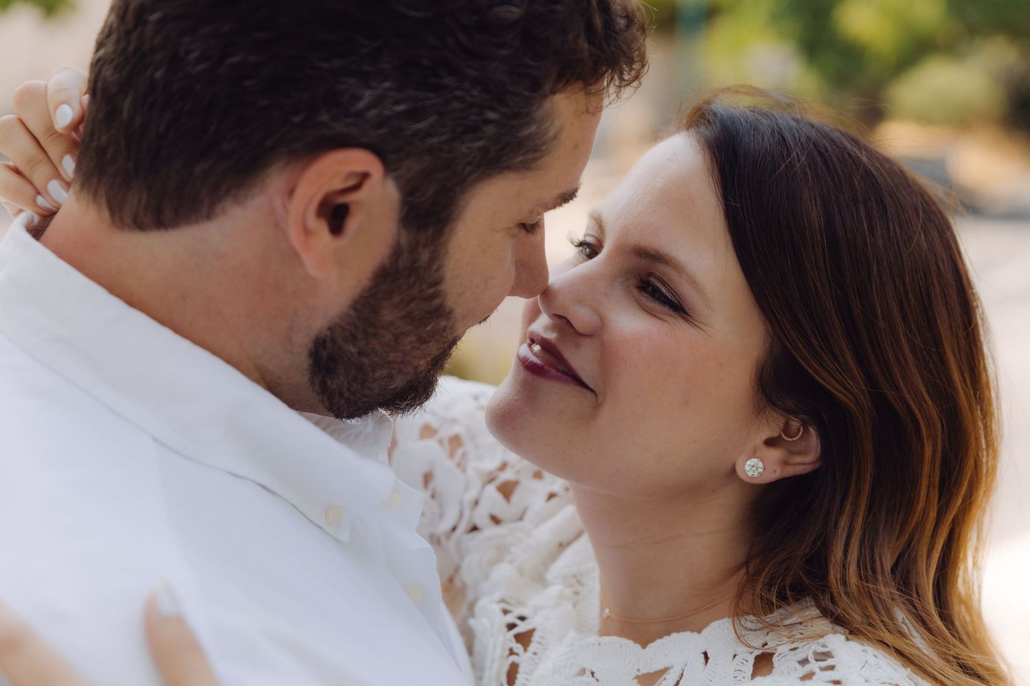 Close-up of couple nearly kissing, woman in white lace dress with pearl earring smiling at bearded man outdoors.