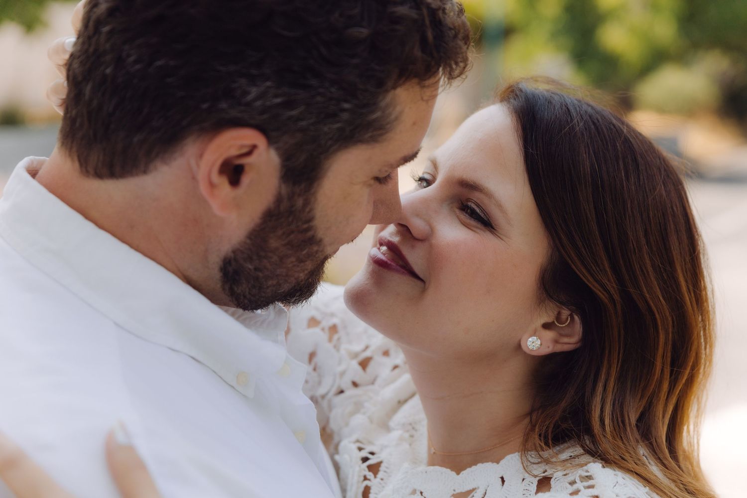 Intimate close-up of smiling couple about to kiss, woman wearing pearl earrings and white lace dress outdoors.