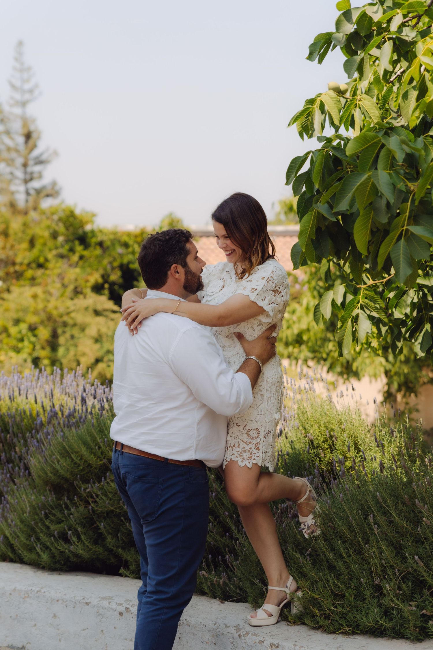 Man in white shirt lifting woman in white lace dress near lavender garden and green trees outdoors.