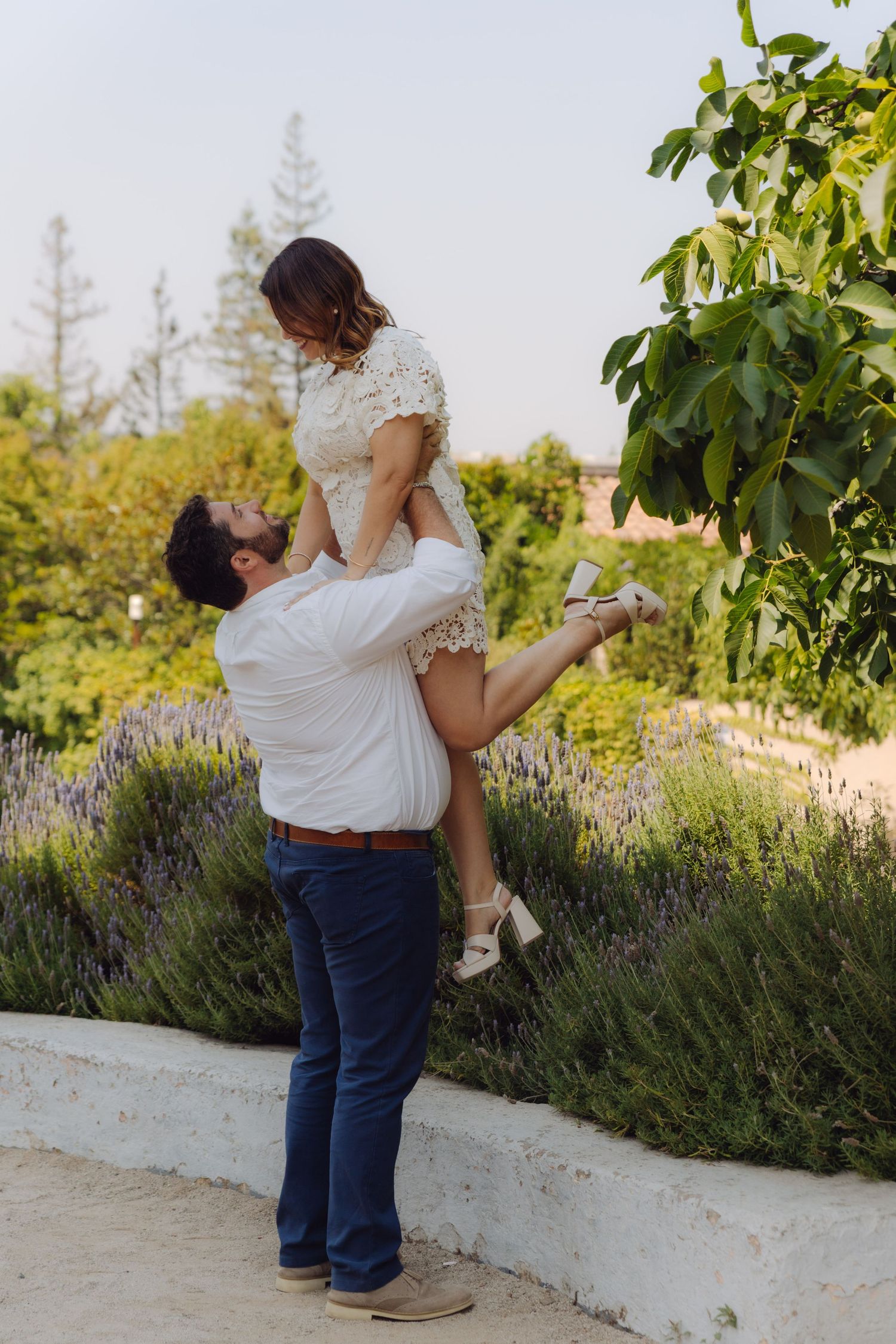 Man in white shirt lifting woman in white lace dress high in the air beside lush green garden and lavender.