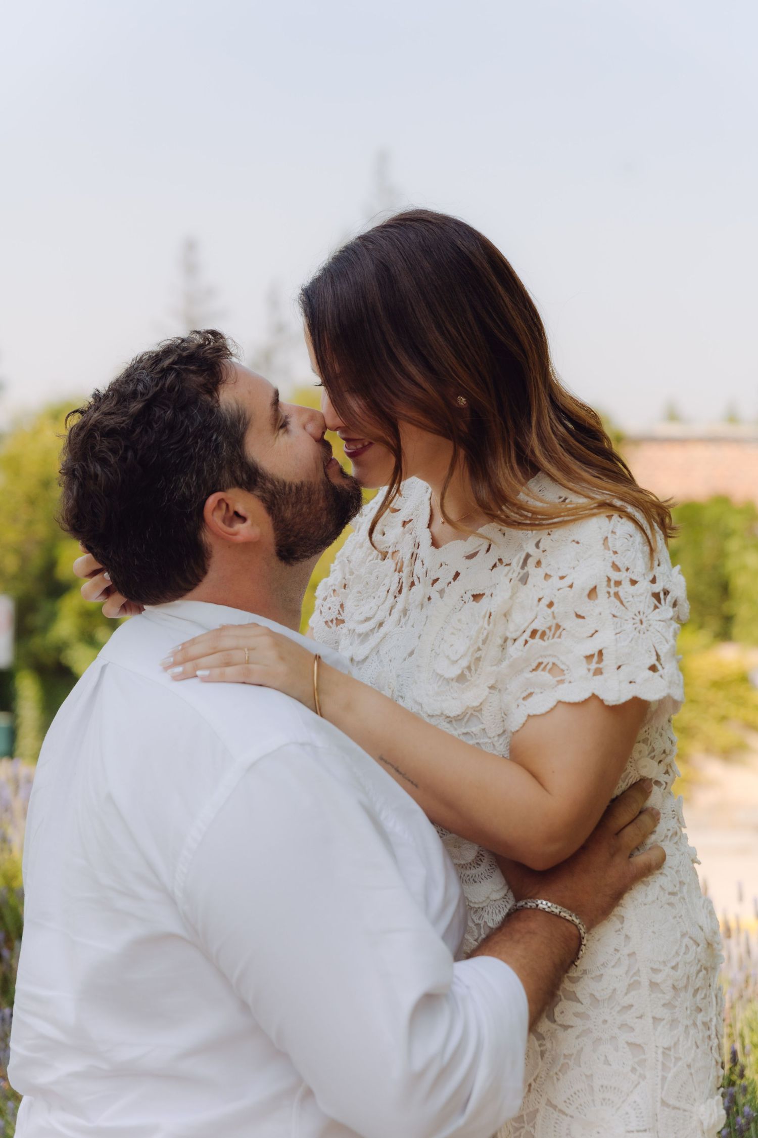 Man in white shirt kissing woman in white lace dress while holding her in a lavender garden outdoors.