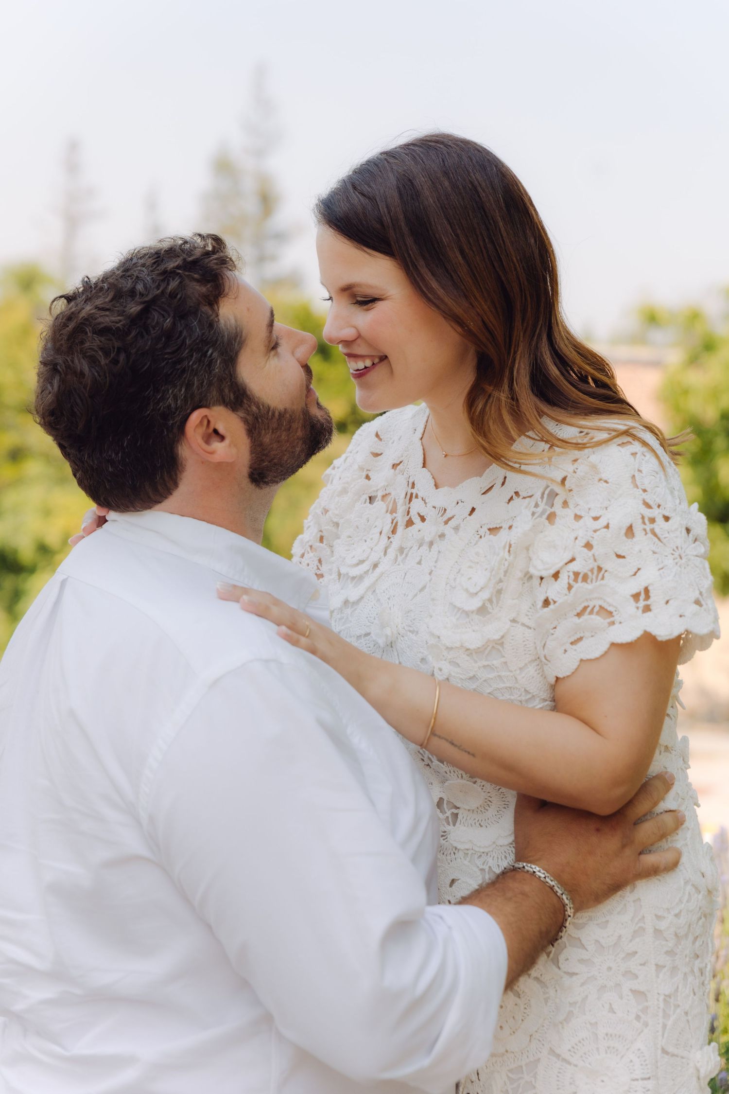 Laughing couple embracing outdoors, woman in white lace dress and bearded man in white shirt in a garden setting.