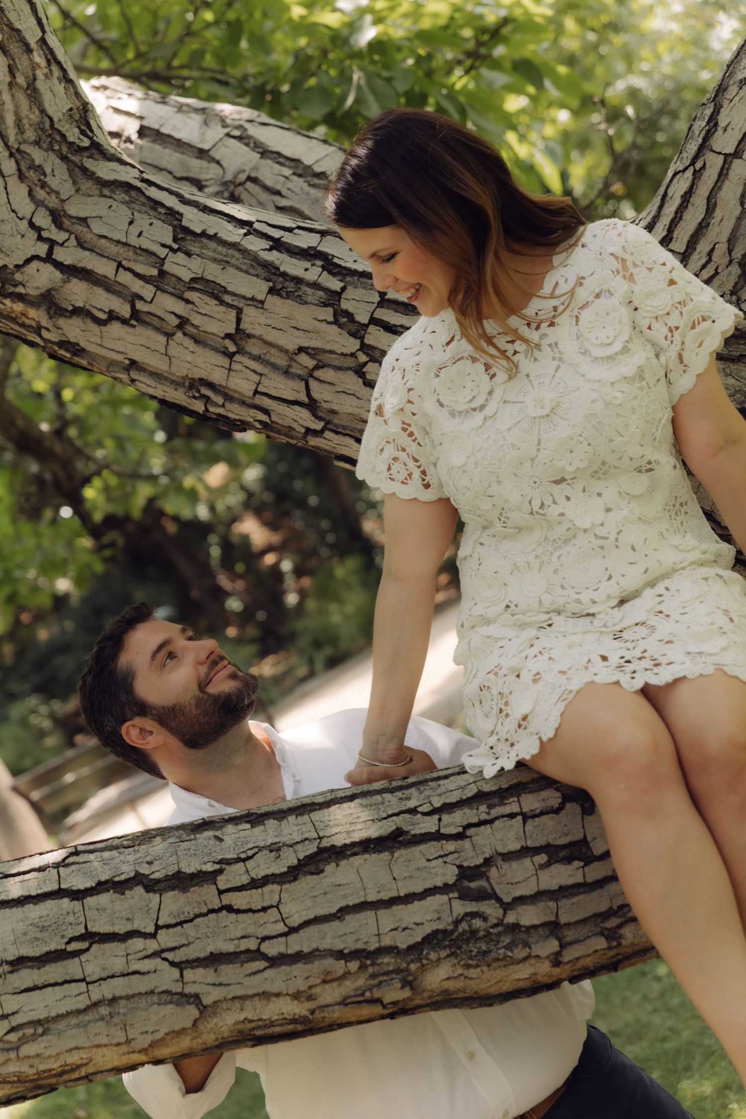 Woman in white lace dress sits on tree branch while bearded man looks up at her in a sunny park.